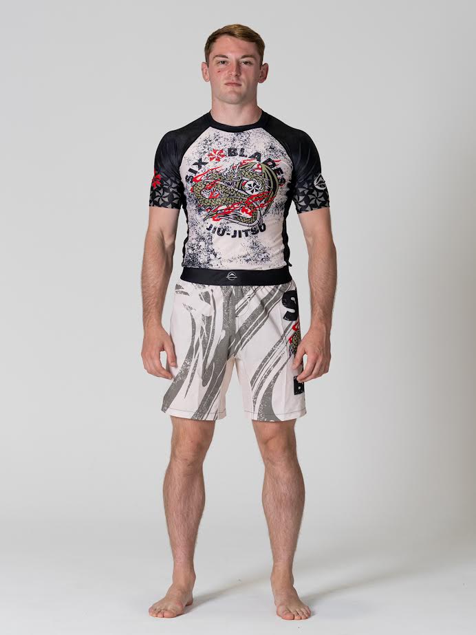 A young man stands barefoot against a plain background, wearing patterned MMA shorts labeled Six Blades Dragon Flex Lite Shorts V2. He faces forward with a neutral expression and relaxed posture.