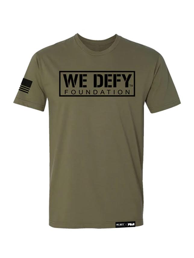 The We Defy Base T-Shirt Military Green features a black WE DEFY FOUNDATION logo on the chest, a black American flag on the right sleeve, and a small label at the bottom hem.