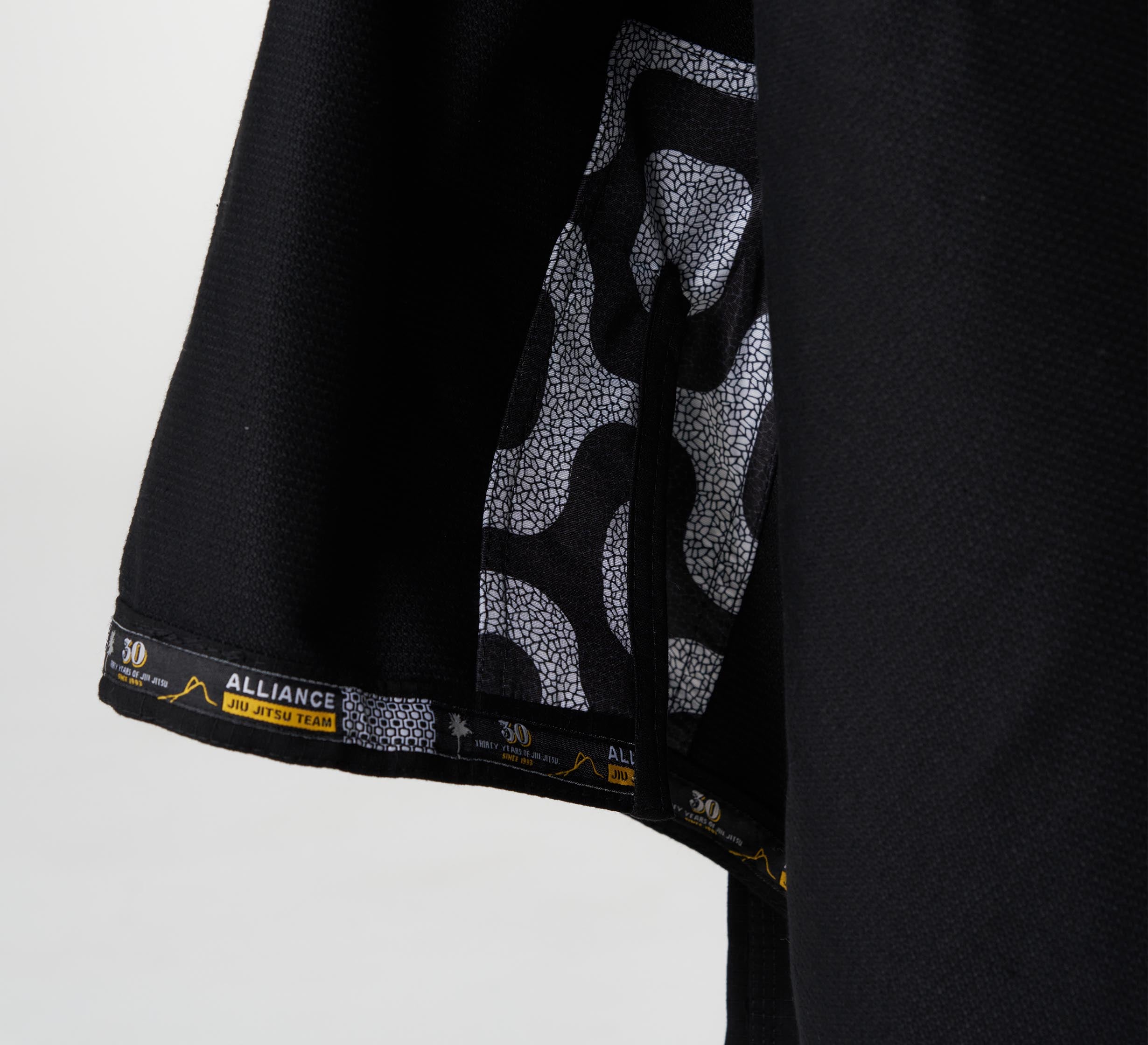 Close-up of the Alliance Limited Edition 30th Anniversary BJJ Gi Black, featuring a patterned black-and-white inner lining and black trim with yellow Alliance Jiu Jitsu Team branding and a mountain graphic at the hem.