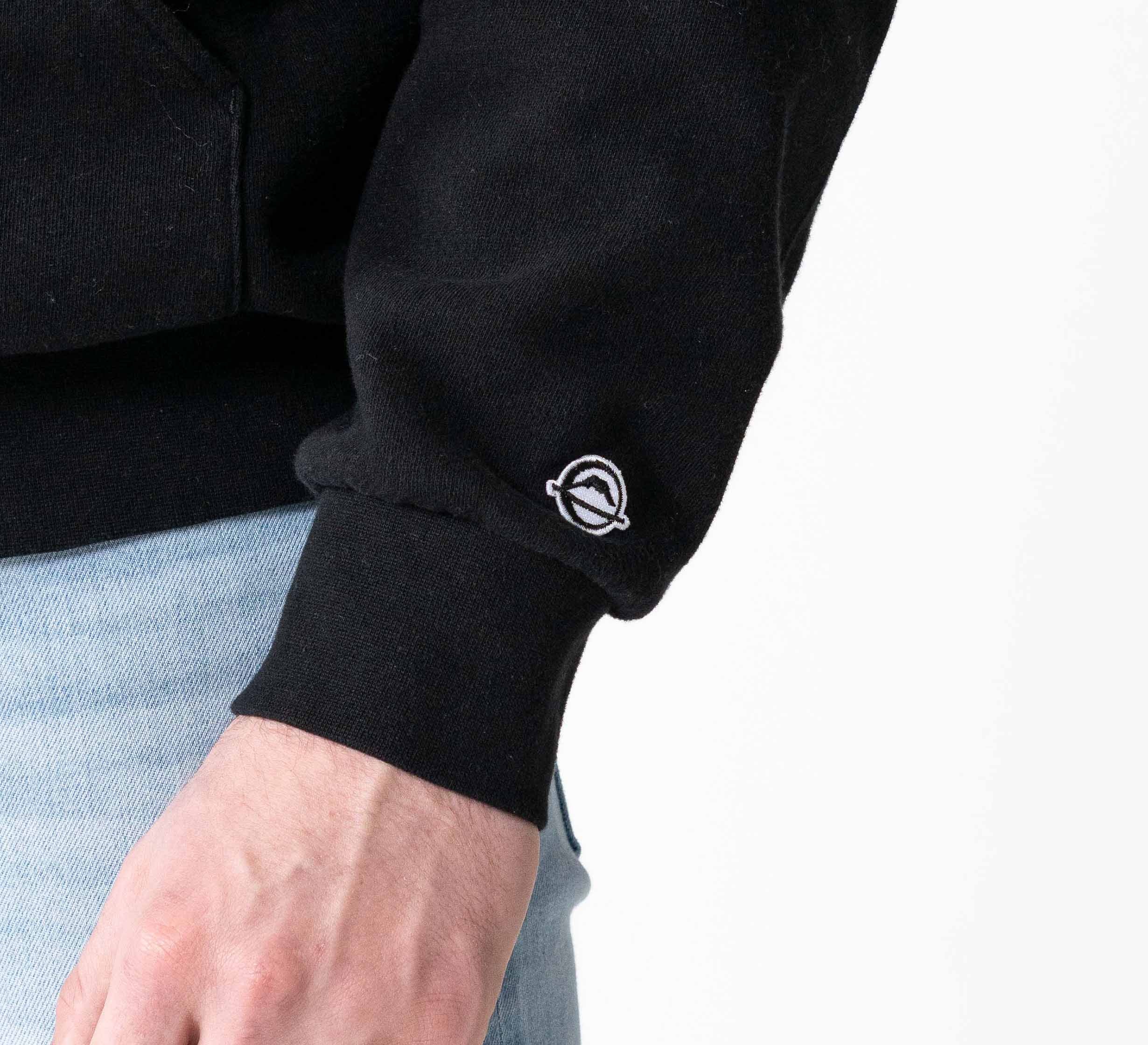 A person wears the Signature Hoodie Black, featuring a small white embroidered planet logo on the sleeve. Only their arm and part of their torso are visible, paired with light blue jeans against a white background.