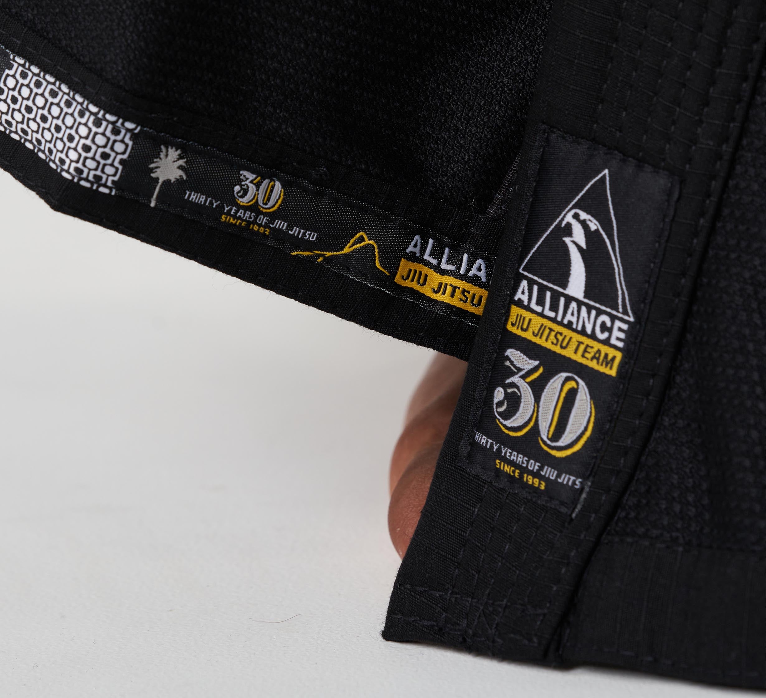 Close-up of the Alliance Limited Edition 30th Anniversary BJJ Gi Black, featuring ALLIANCE JIU JITSU TEAM and 30 logos marking thirty years since 1993. A hand holds the gi’s edge, highlighting its detailed stitching and commemorative patches.