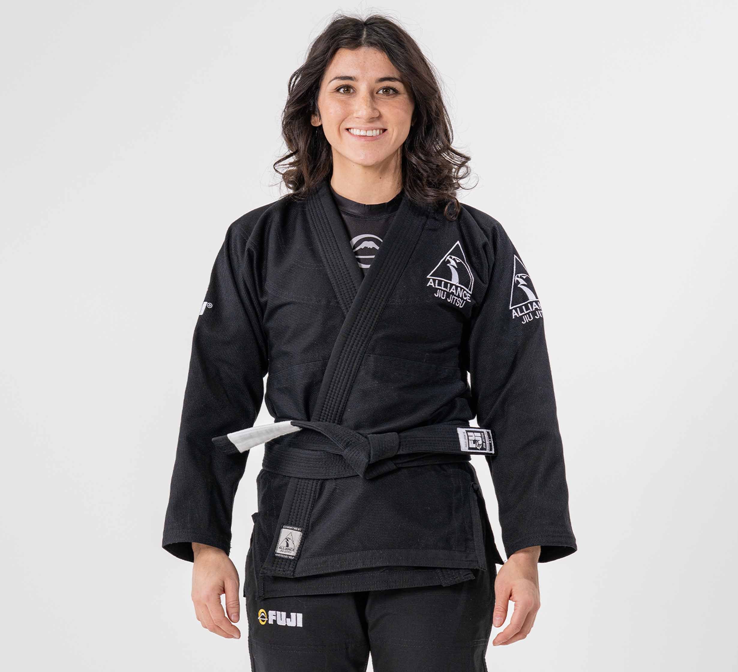 A woman with long dark hair smiles, wearing the Womens FUJI x Alliance Standard BJJ Gi Black with a white belt. She stands against a plain light background, and Alliance logos are visible on the chest and sleeve of the gi.