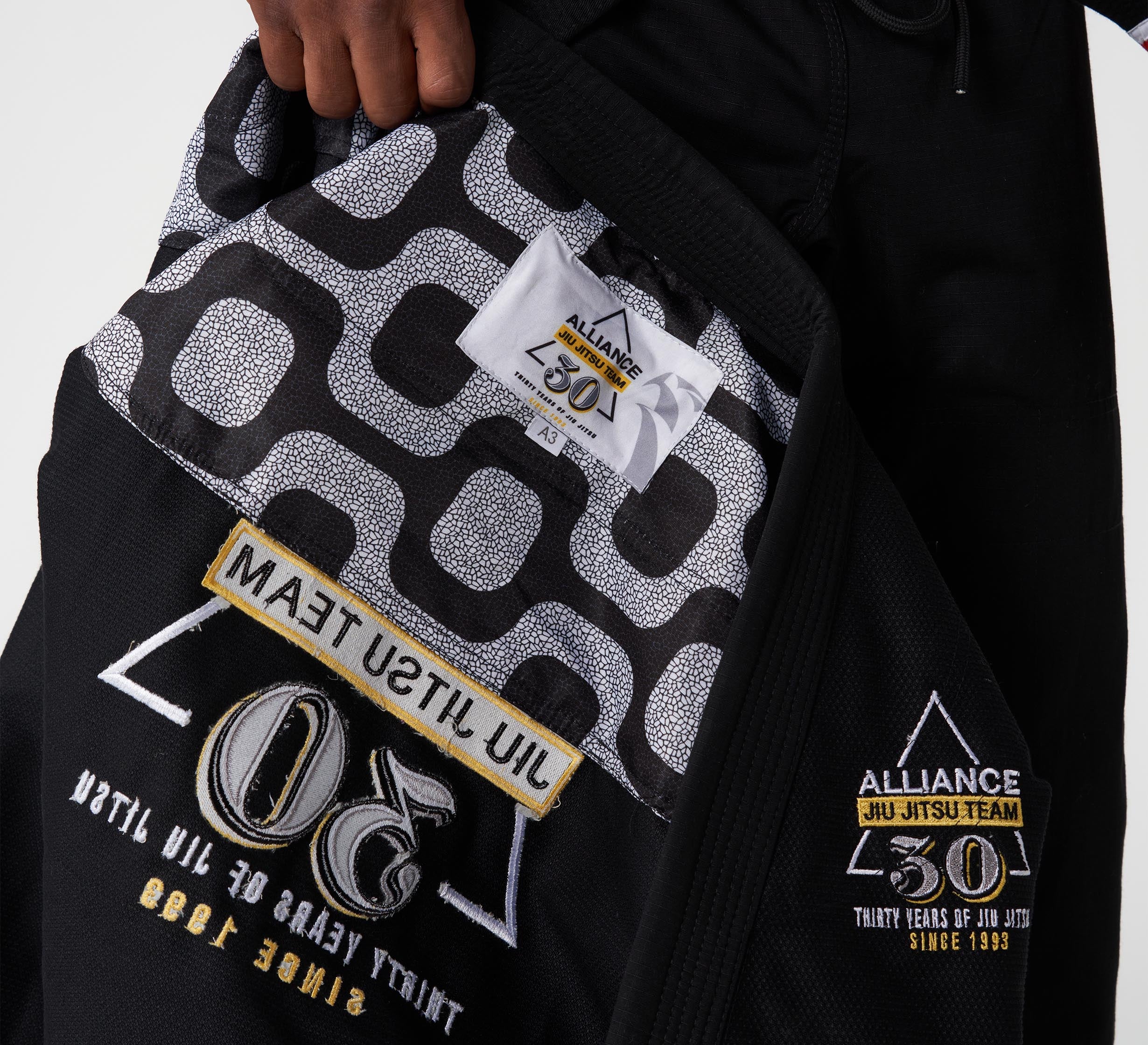 A close-up of the Alliance Limited Edition 30th Anniversary BJJ Gi Black, featuring gold and white embroidered patches and a black-and-white patterned inner lining. A hand holds the jacket open to display its details.