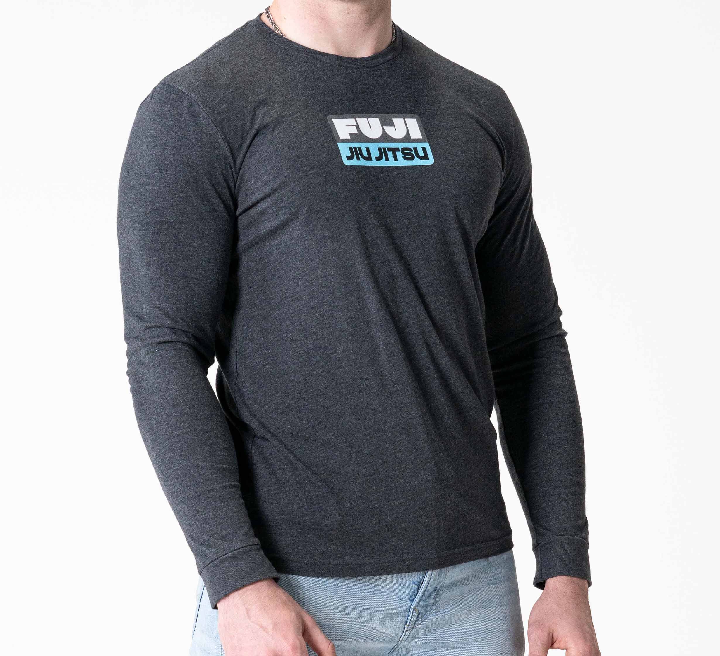A person wears the Retro Long Sleeve Shirt Charcoal, featuring FUJI JIU JITSU in blue and white on the front, paired with light blue jeans and standing against a plain white background.