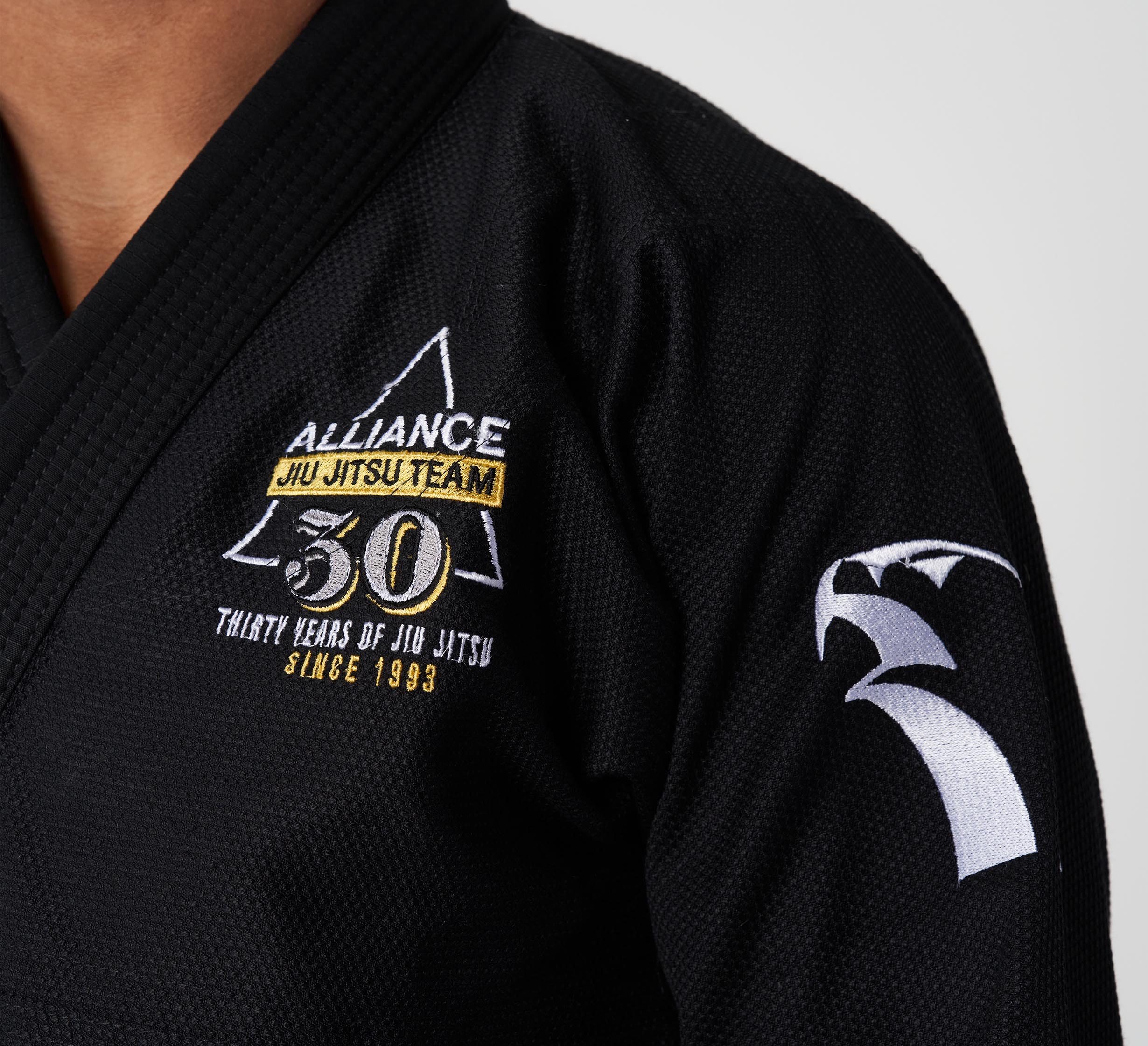 Close-up of a person wearing the Alliance Limited Edition 30th Anniversary BJJ Gi Black, featuring embroidered Alliance Jiu Jitsu Team patches, "30 Years of Jiu Jitsu Since 1993," and a white shark fin design on the sleeve.