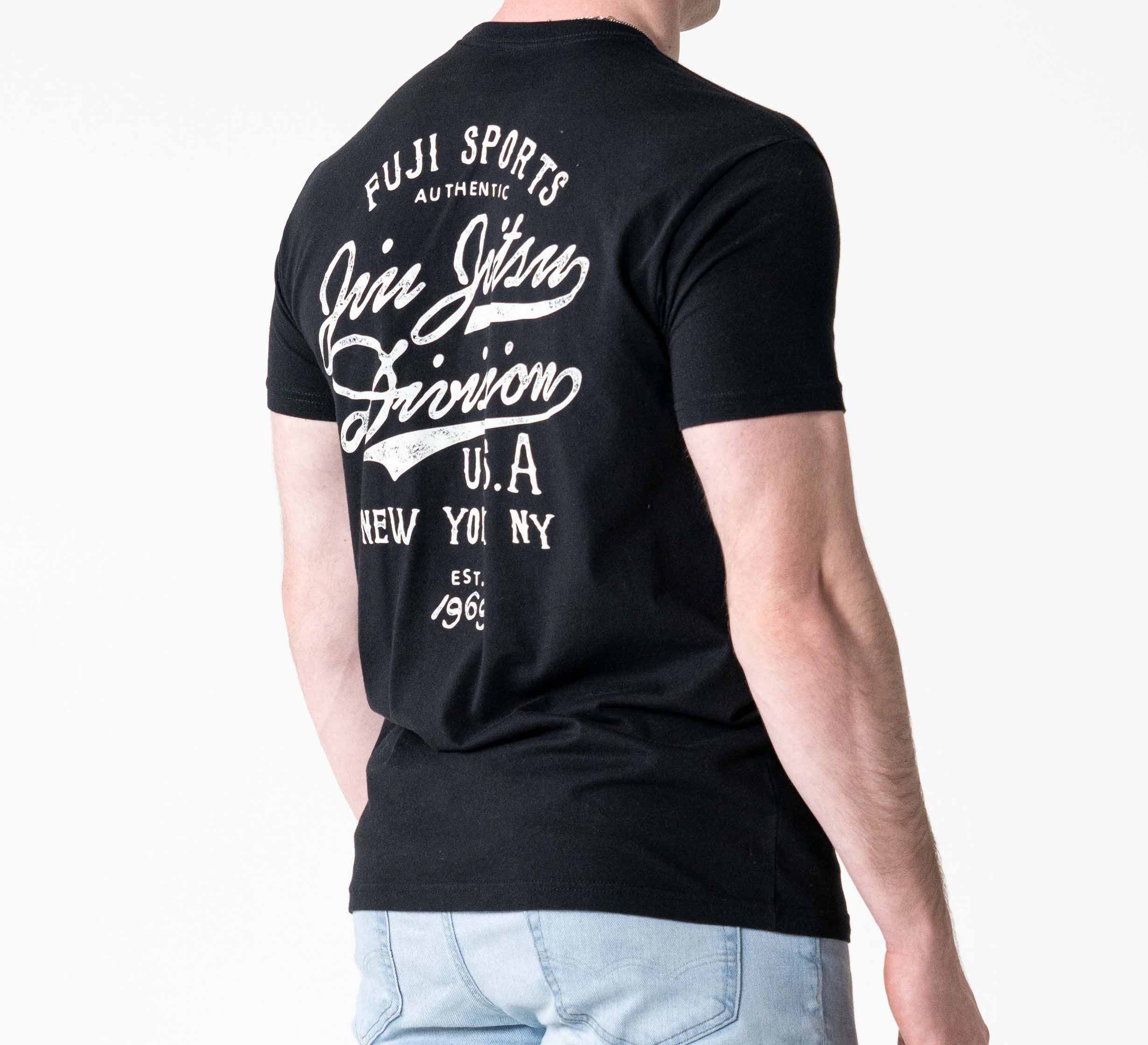 A person wearing the Jiu Jitsu Flow T-Shirt Black, featuring white scripted text on the back: "Fuji Sports Authentic Jiu Jitsu Division USA New York Est. 1969," is facing away and paired with light blue jeans.