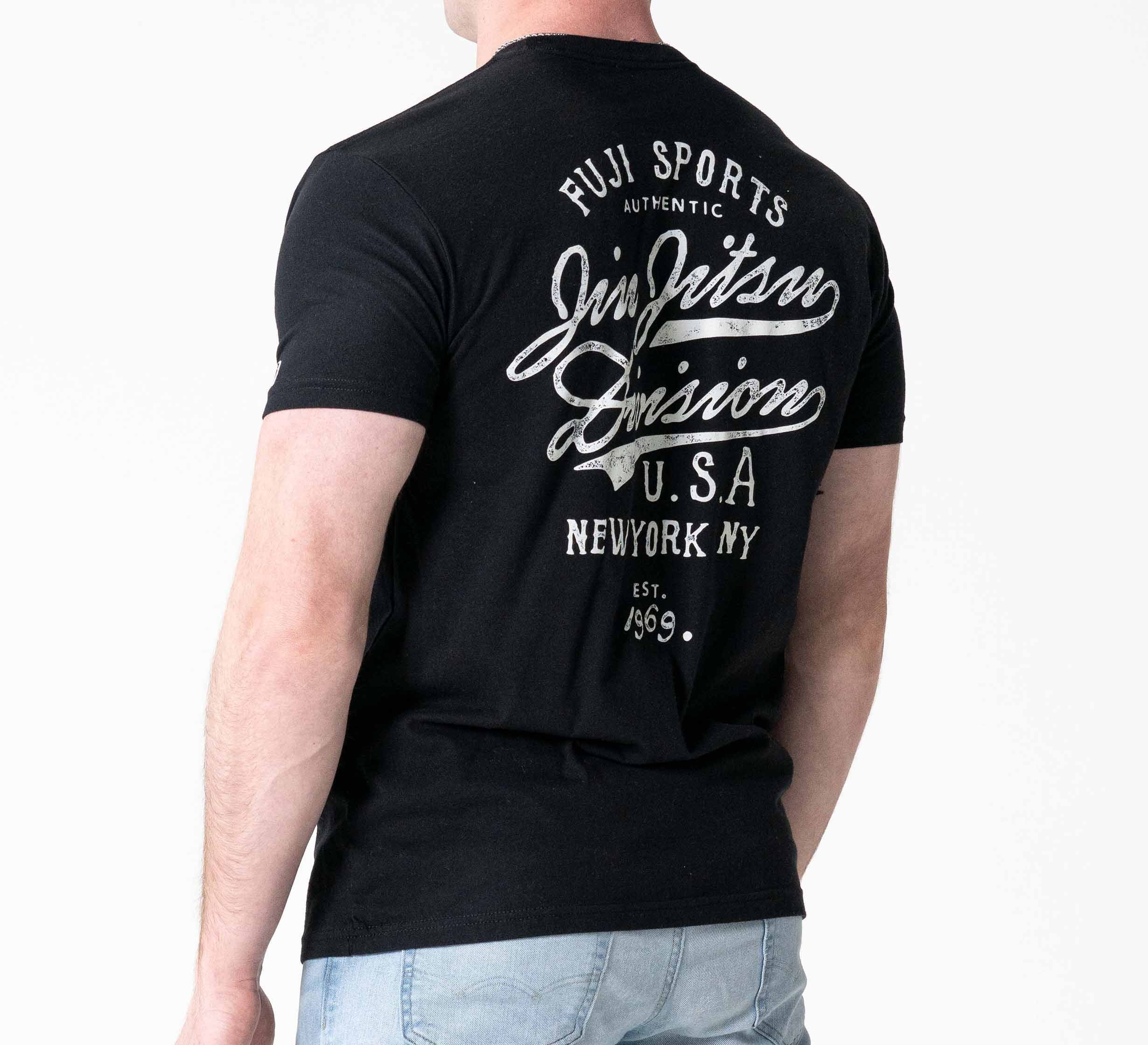 A person faces away from the camera, wearing the Jiu Jitsu Flow T-Shirt Black with white “Fuji Sports Jiu Jitsu Division U.S.A. New York NY Est. 1969” text on the back, paired with light blue jeans.