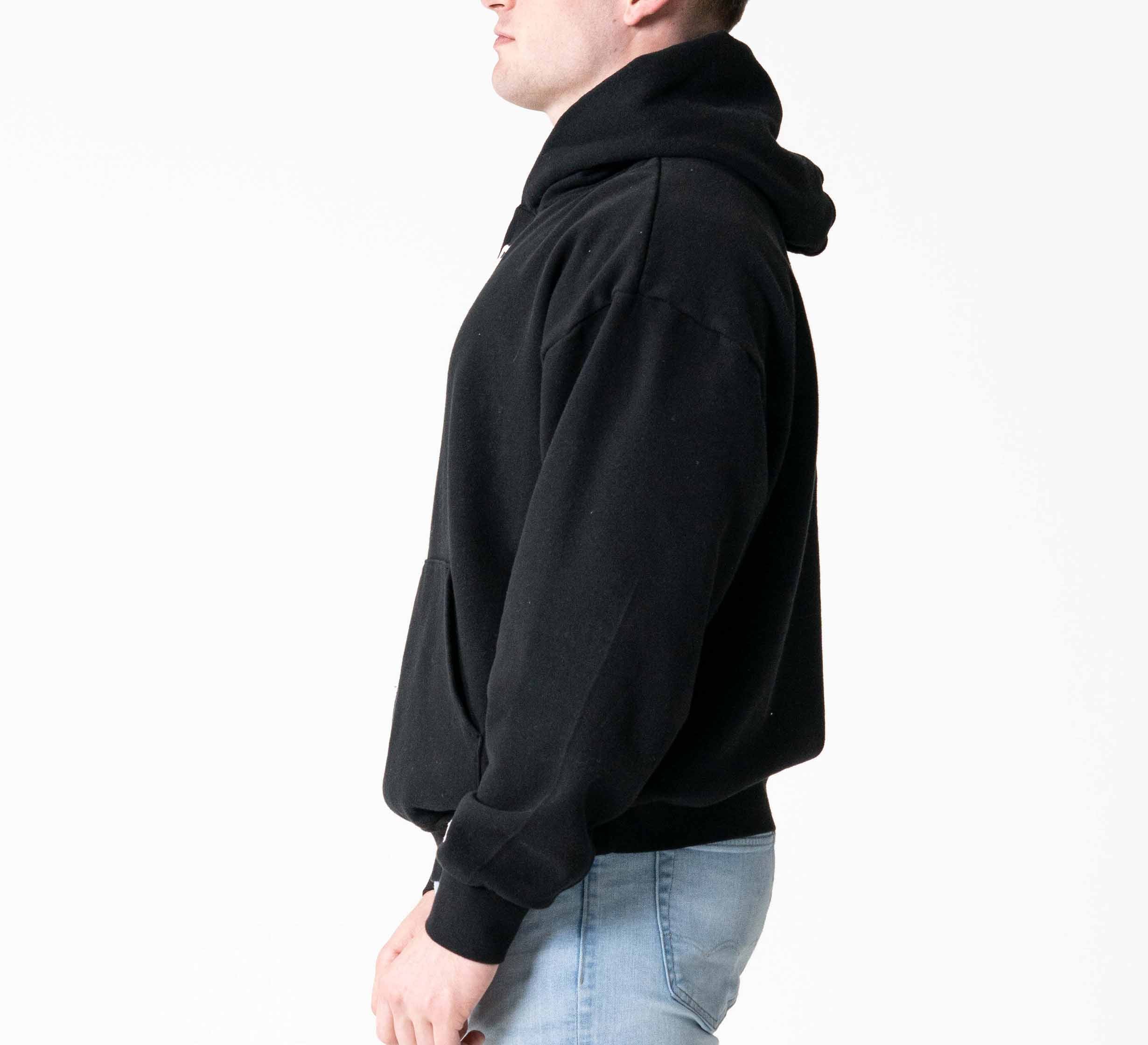 A person stands in profile against a plain white background wearing the Signature Hoodie Black and light blue jeans.