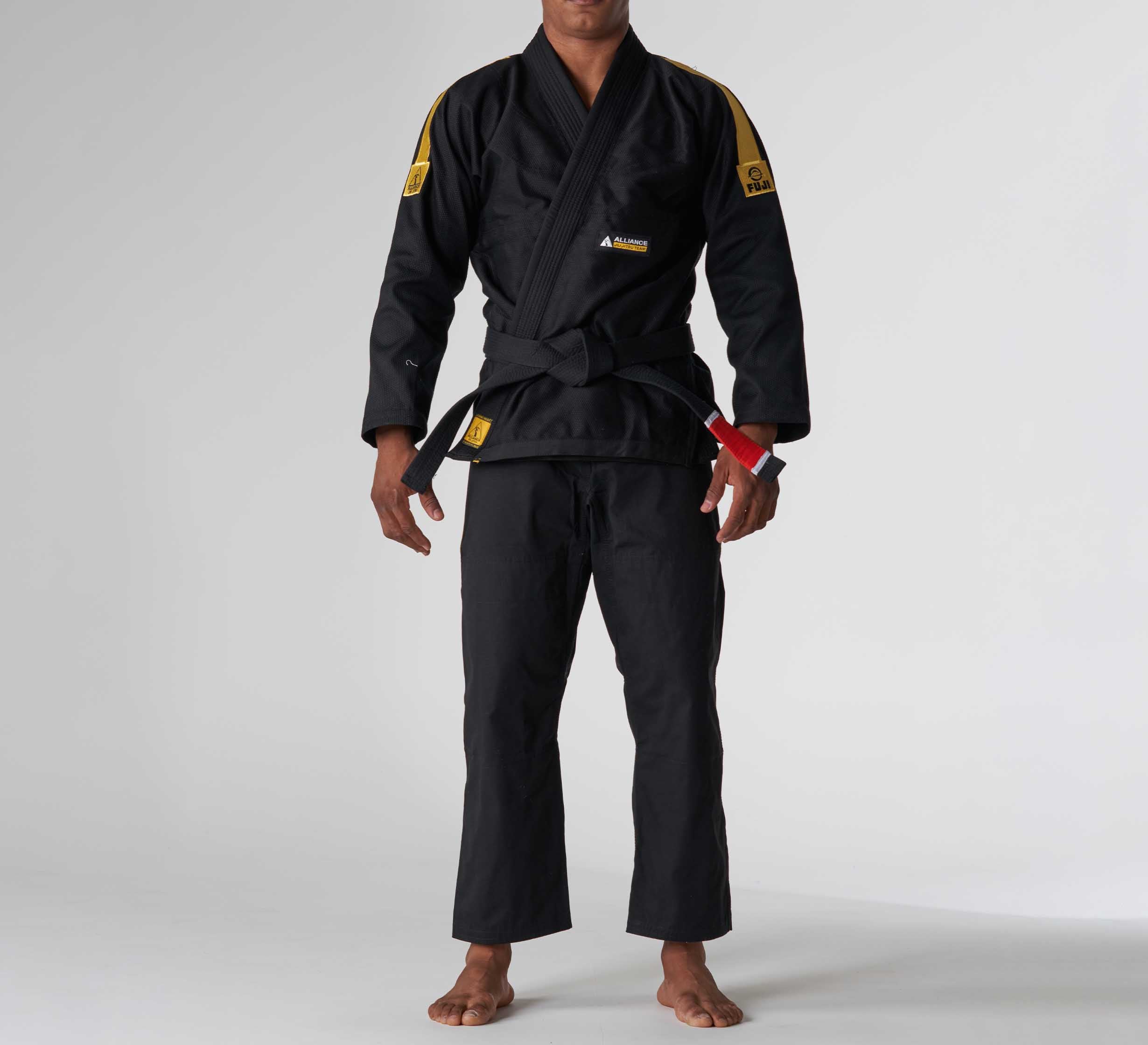 A person stands barefoot on a plain background wearing the FUJI x Alliance Sekai BJJ Gi Black with yellow patches and a black belt. Their arms are relaxed at their sides, and their face is not visible.
