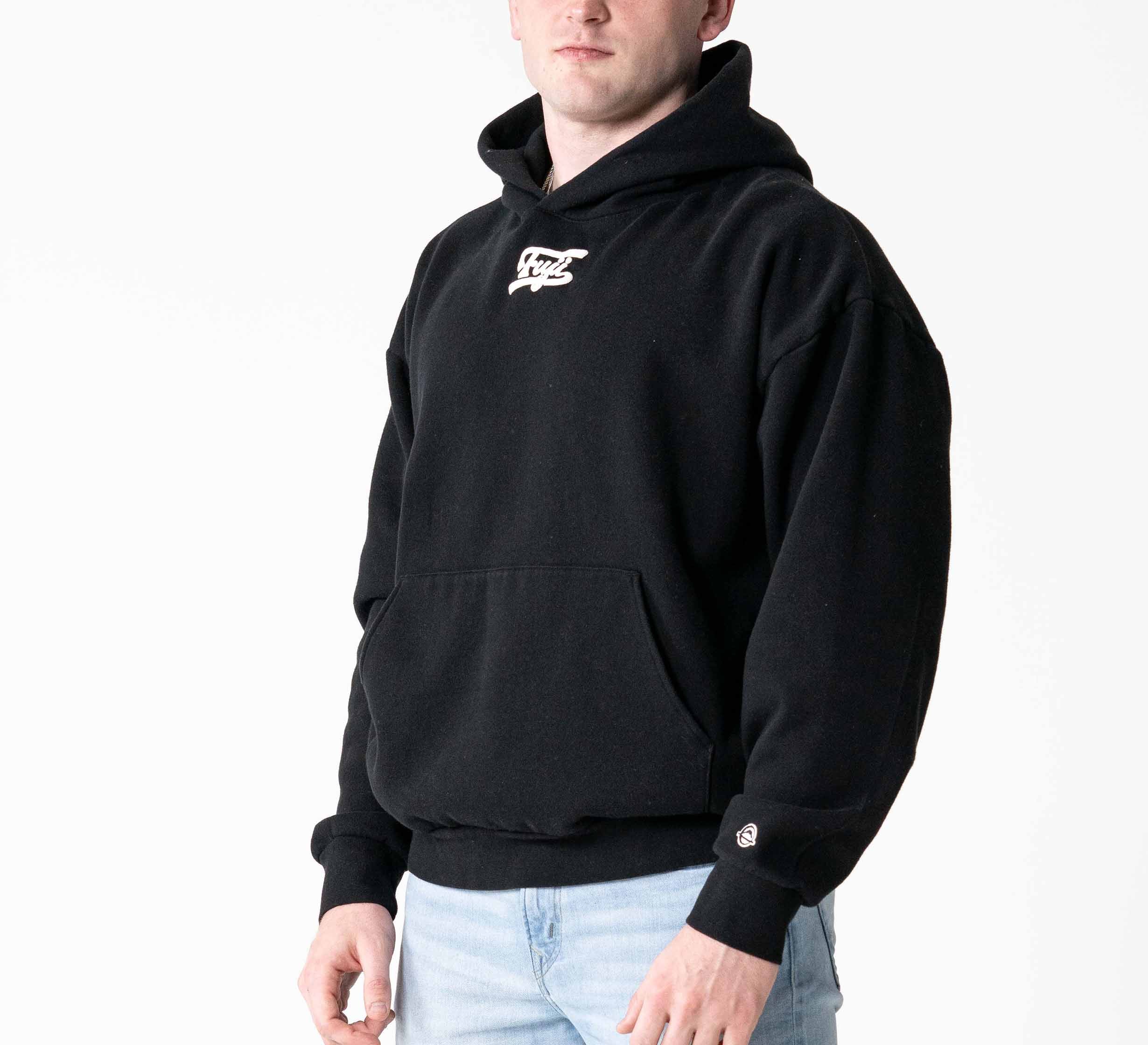 A person wearing the Signature Hoodie Black, featuring a small white logo on the chest, paired with light blue jeans and standing against a plain white background.