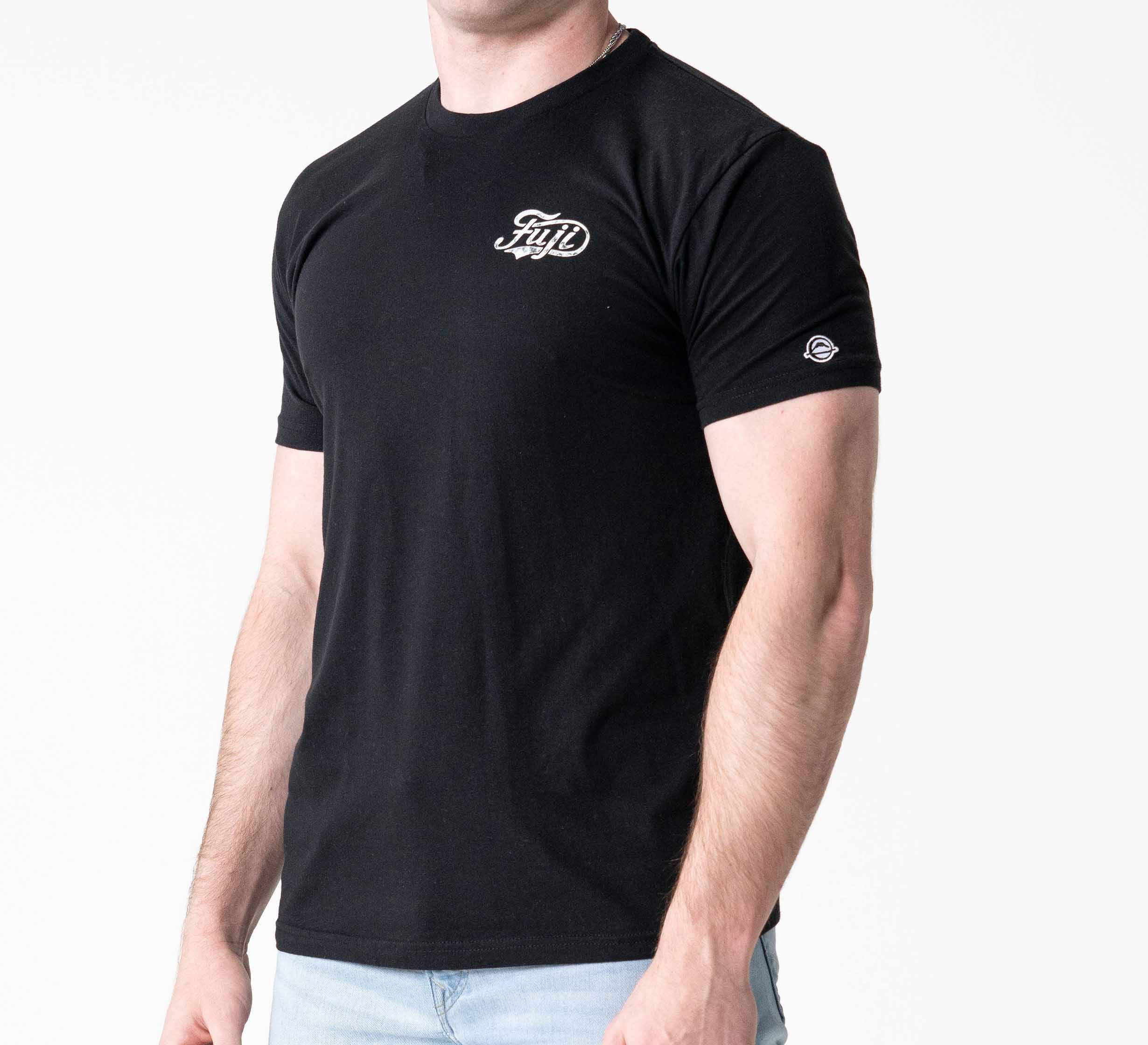 Someone wears the Jiu Jitsu Flow T-Shirt Black, a fitted black short-sleeve tee featuring a small white chest logo and sleeve detail, paired with light blue jeans against a plain white background.