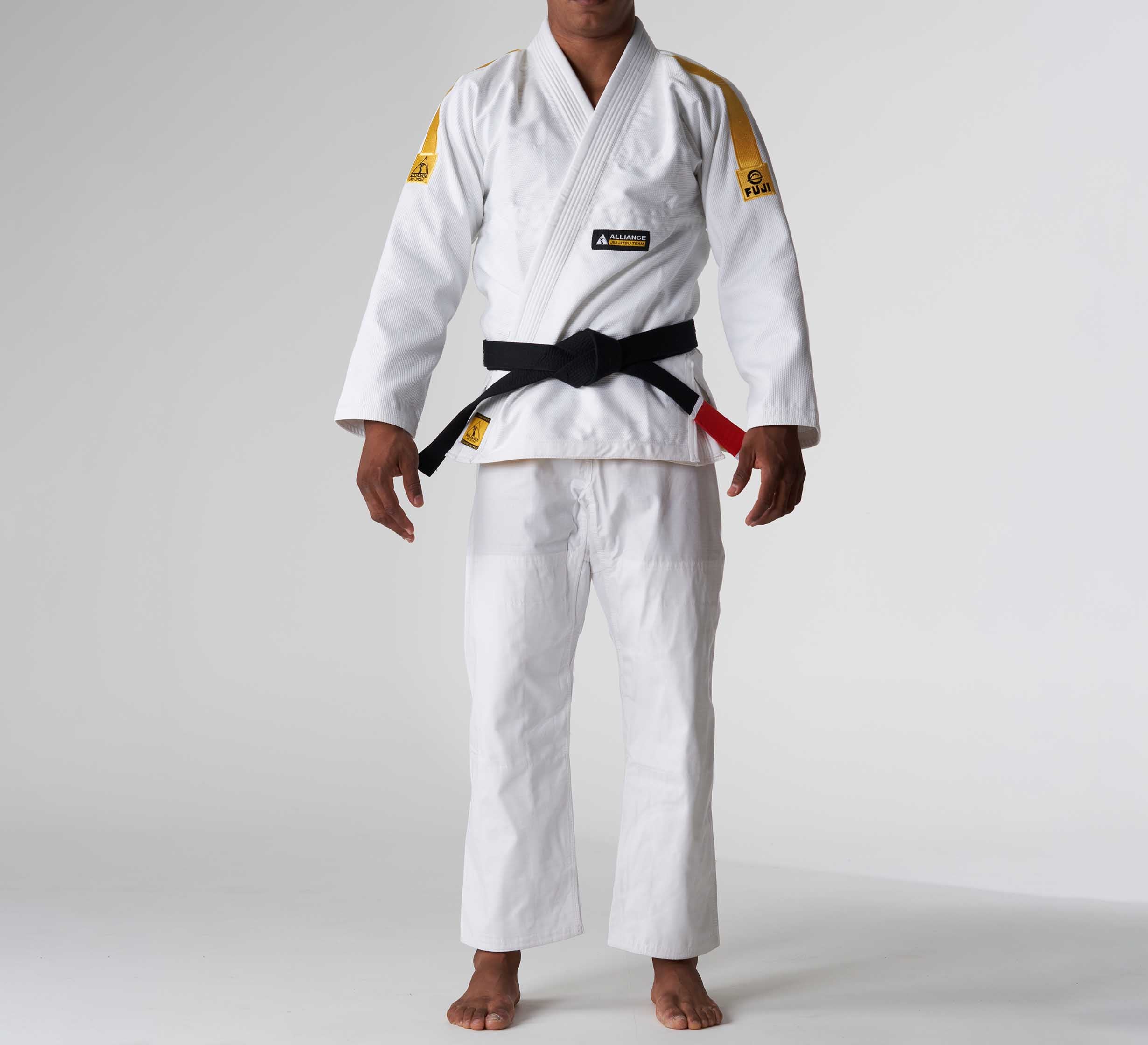 Someone stands barefoot against a plain, light background wearing the FUJI x Alliance Sekai BJJ Gi White with a black belt; their face is not visible.