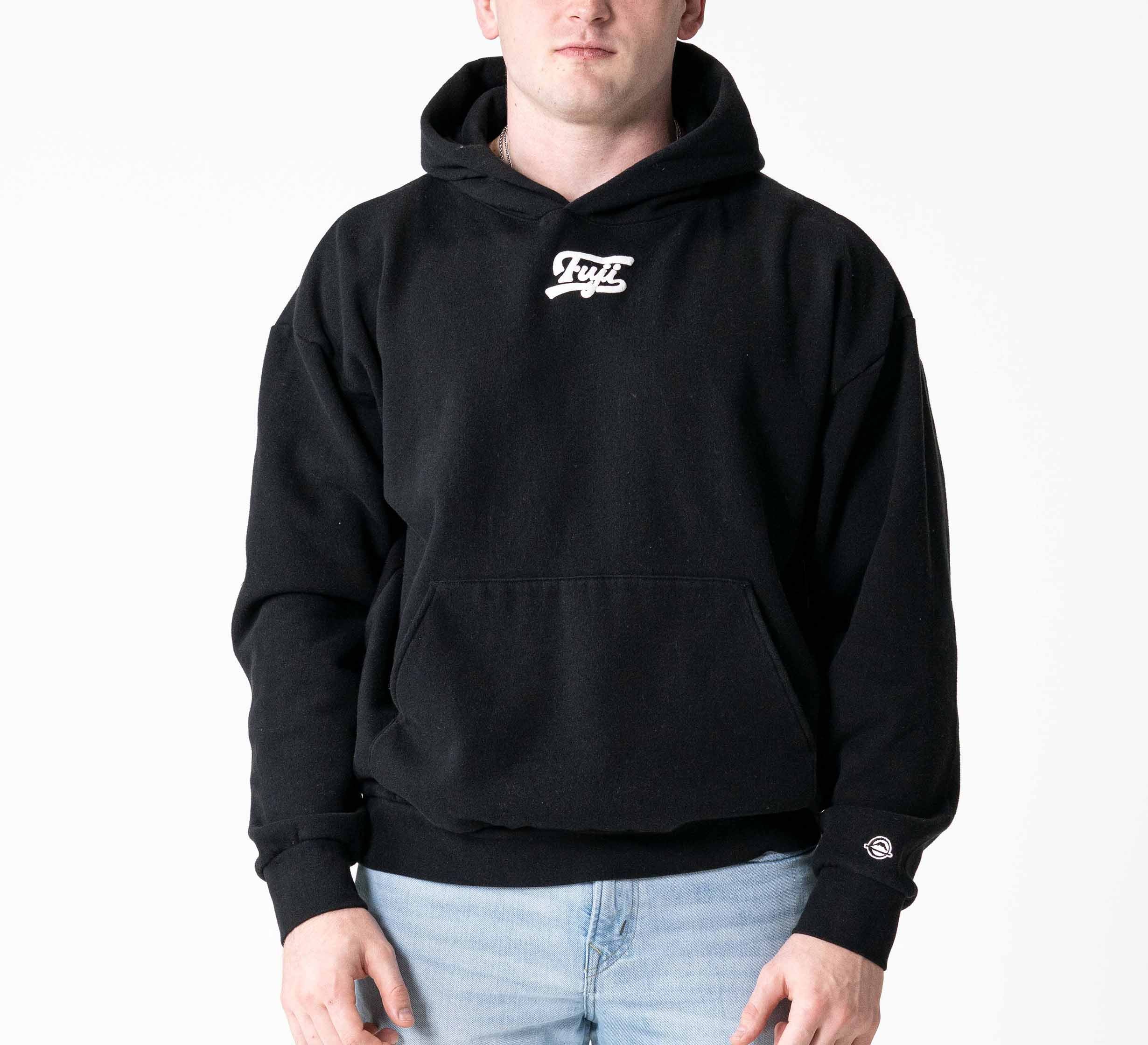 A person wears the Signature Hoodie Black, featuring a small white chest logo and a front pocket, paired with light blue jeans, standing against a plain white background with hands relaxed by their sides.