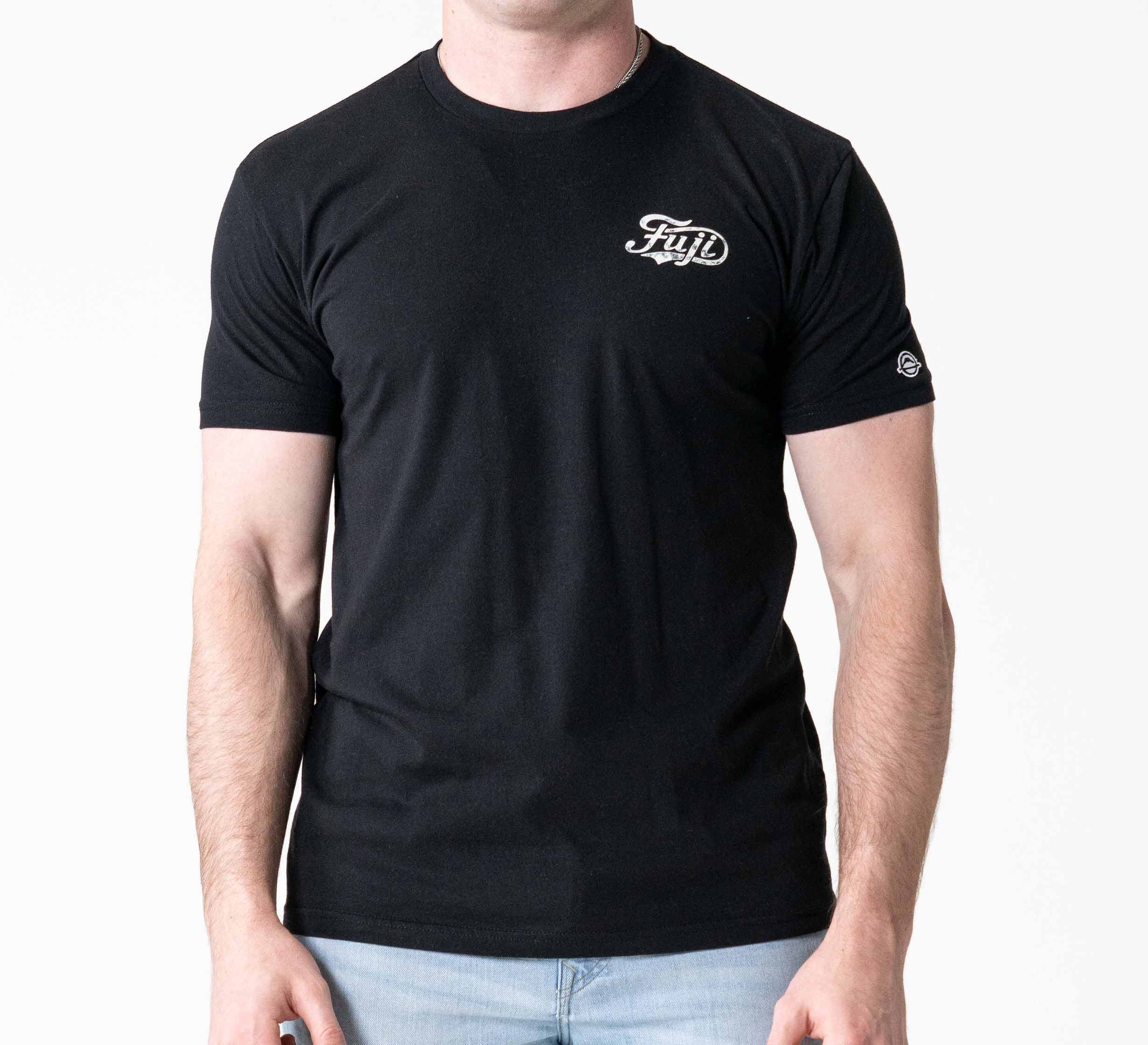 A person wearing the Jiu Jitsu Flow T-Shirt Black with "Fiji" in white on the left chest and a small white logo on the left sleeve, paired with light blue jeans against a plain white background.