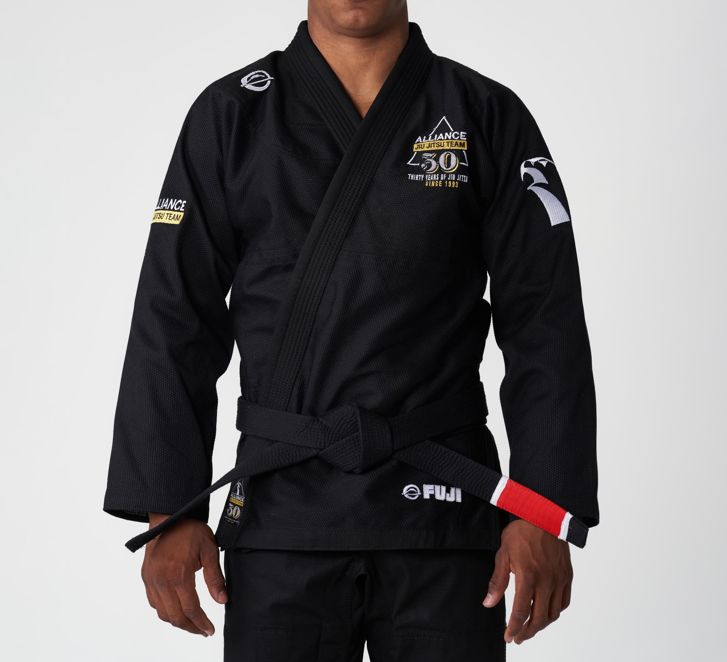 A person wearing the Alliance Limited Edition 30th Anniversary BJJ Gi Black with Alliance 30 Years and Fuji patches, and a black belt with red and white bars, stands against a plain white background.