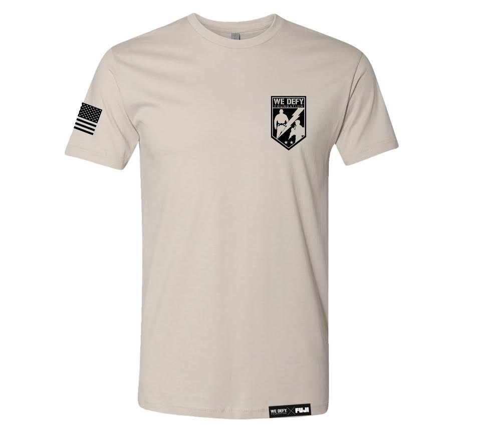 The We Defy Core T-Shirt Sand is a beige tee with a black American flag on the right sleeve, a WE DEFY crest on the left chest, and two small rectangular tags at the bottom front hem.