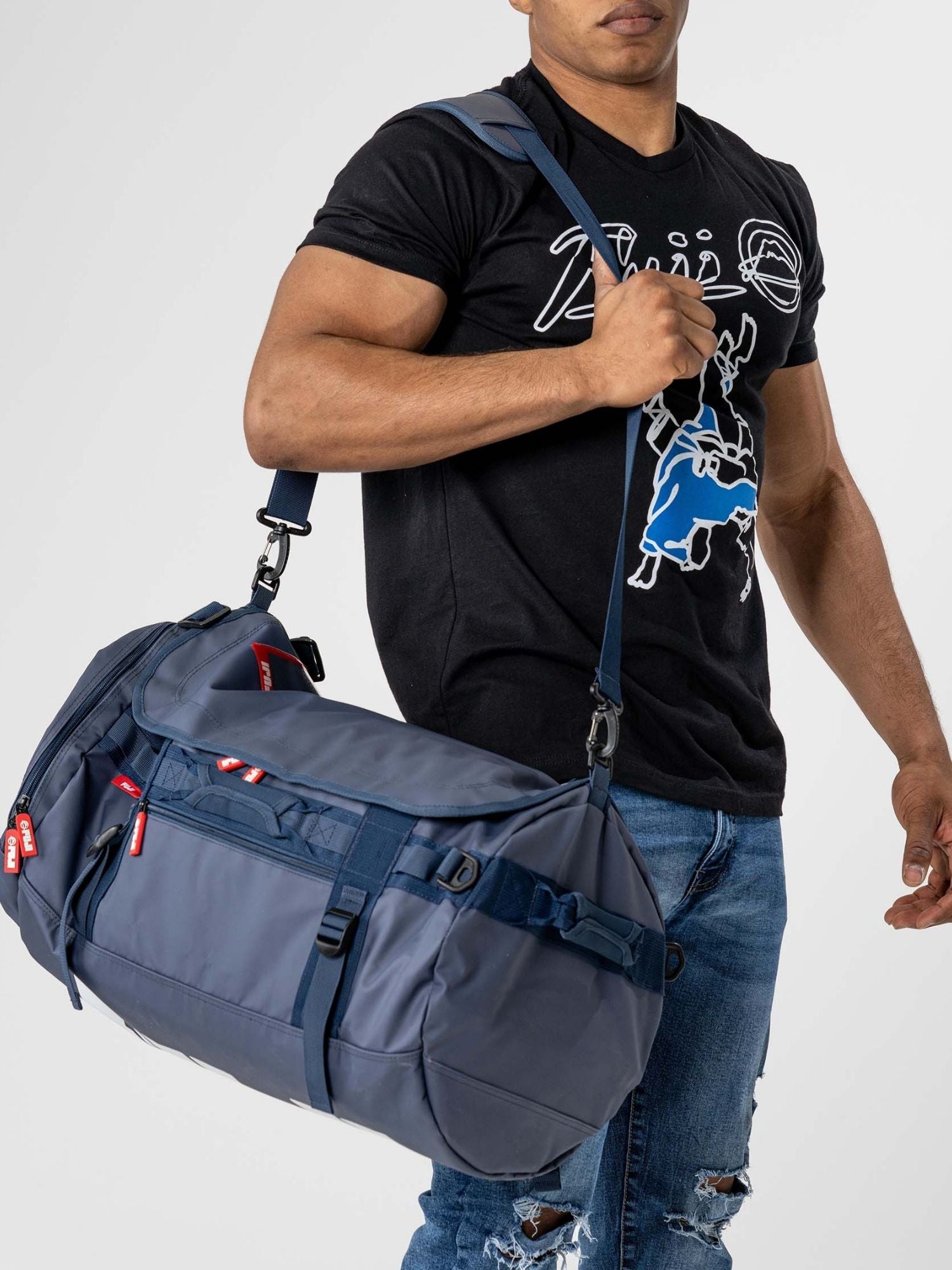 A man in a black graphic tee and ripped blue jeans carries the Comp Convertible Backpack Duffle Navy over his shoulder. The plain, light-colored background highlights the bag's sleek design.