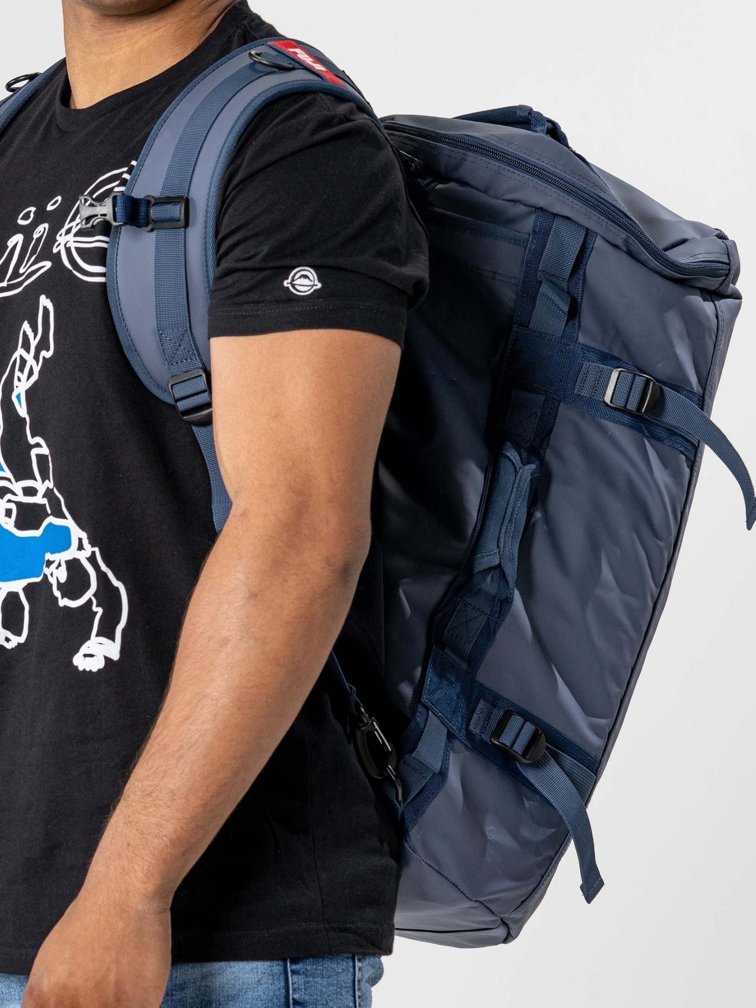 A person in a black graphic tee and blue jeans carries the Comp Convertible Backpack Duffle Navy by its shoulder straps like a backpack. The cropped image shows only their torso and the bag.