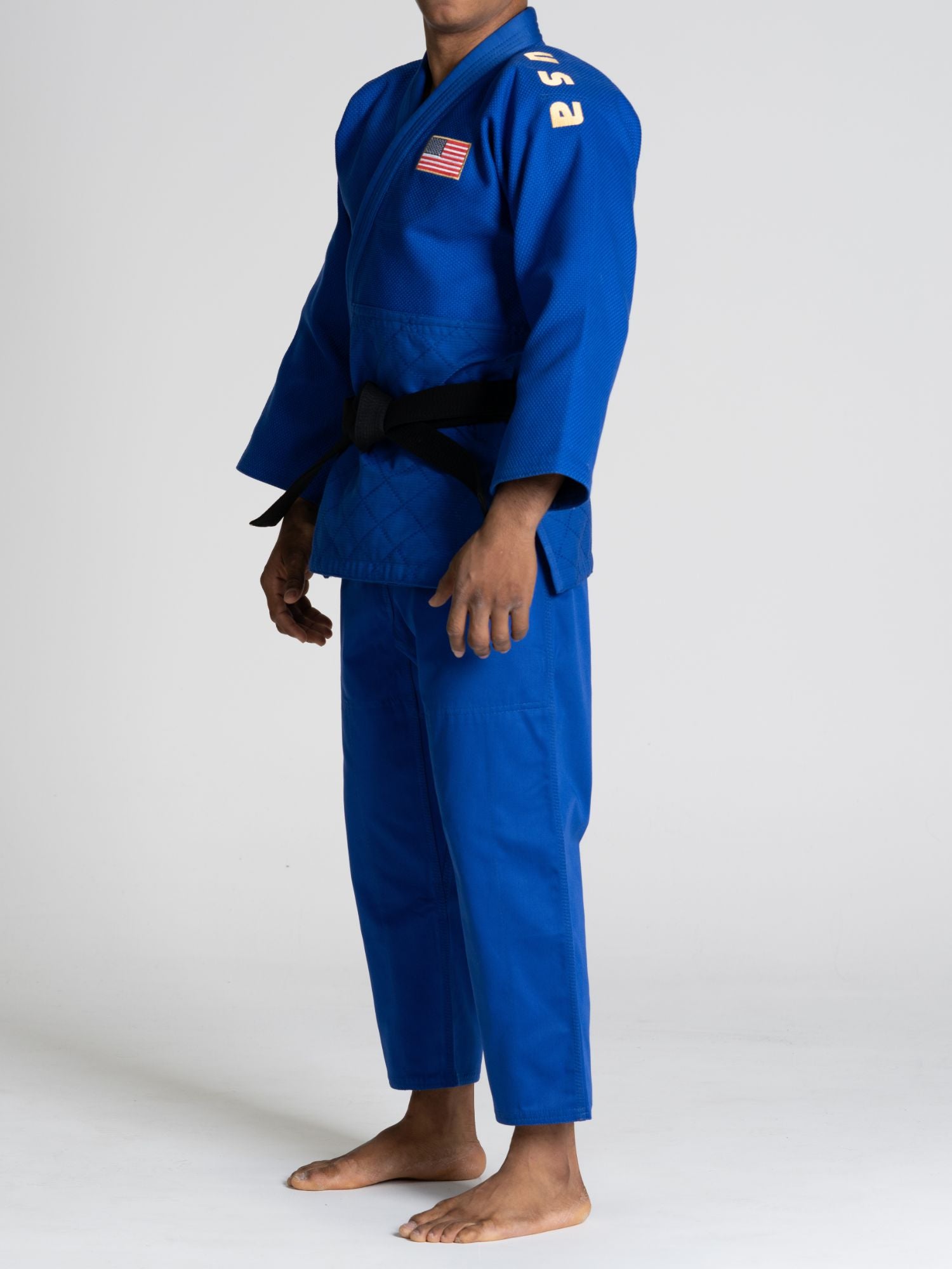 A person in the USA Judo Euro Competition Judo Gi Blue with a black belt stands barefoot on a white background. An American flag patch and a yellow number are visible on the uniform’s shoulder.