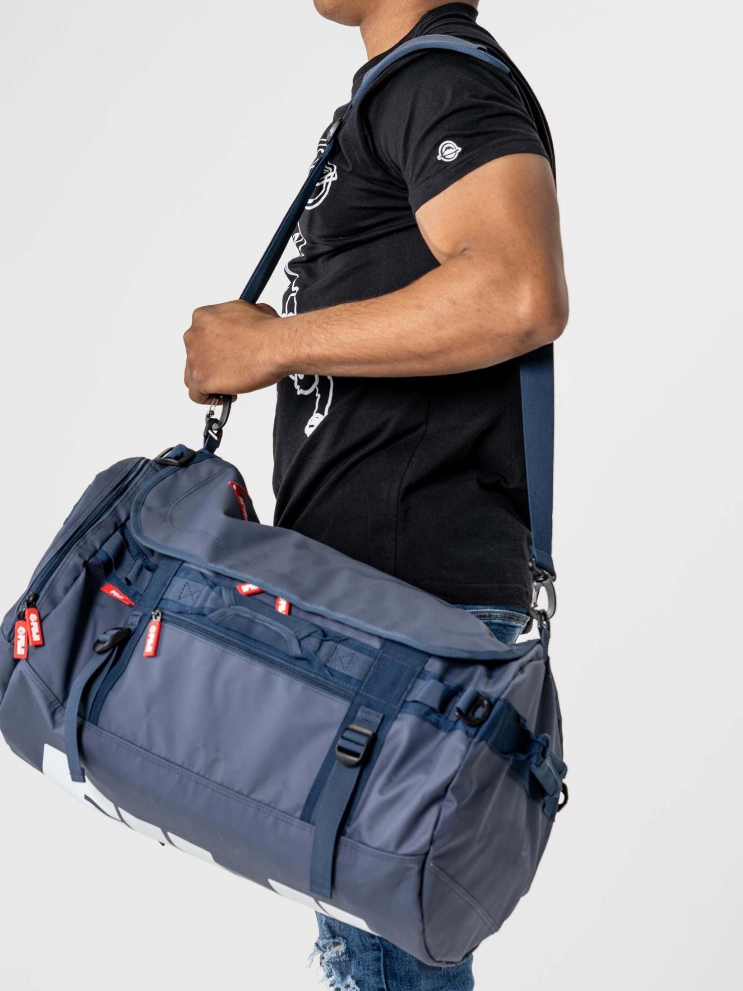 A person in a black t-shirt and blue jeans carries the Comp Convertible Backpack Duffle Navy, featuring red zipper pulls, multiple pockets, a shoulder strap, and a large size. The background is plain and light-colored.