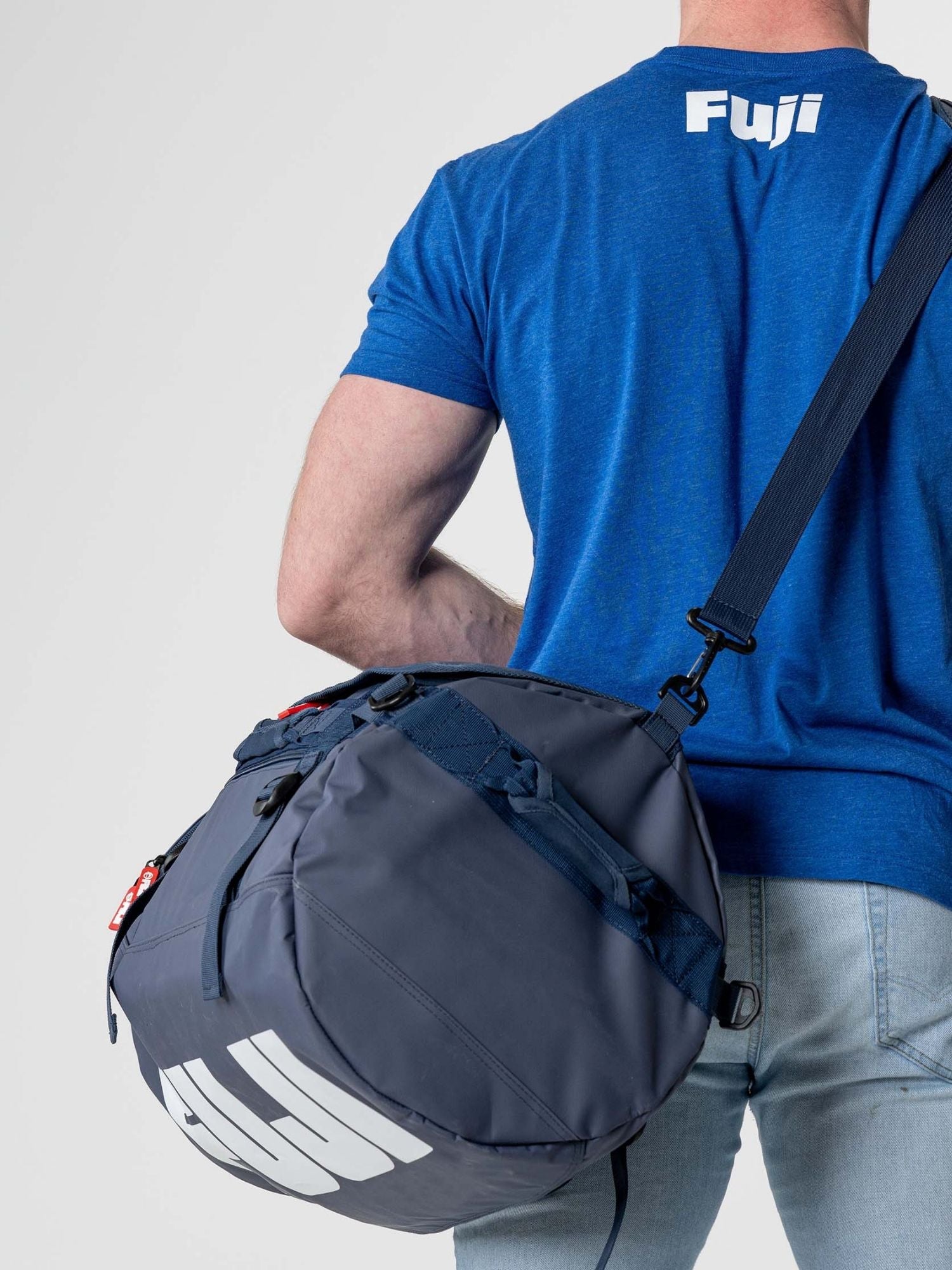 A person wearing a blue Fuji t-shirt and light jeans carries the Comp Convertible Backpack Duffle Navy, a large round navy bag with "Fuji" in white on the strap and side, slung over their shoulder.