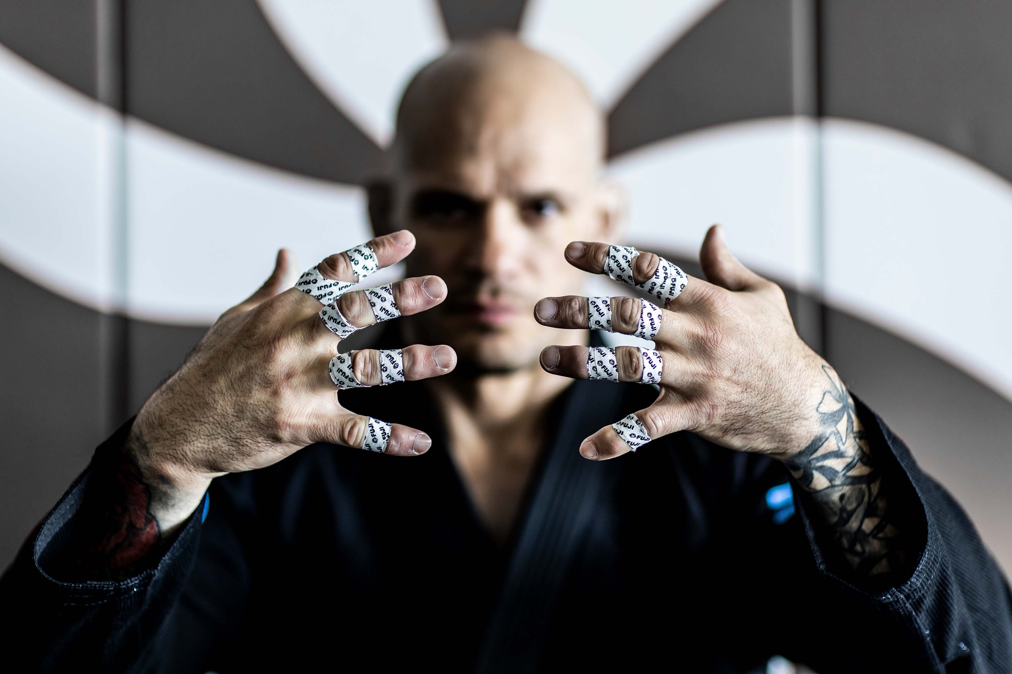 A person in a martial arts gi shows both hands forward, each finger taped for support. The background features bold white geometric patterns. The focus is on the hands and finger tape, not the persons face.