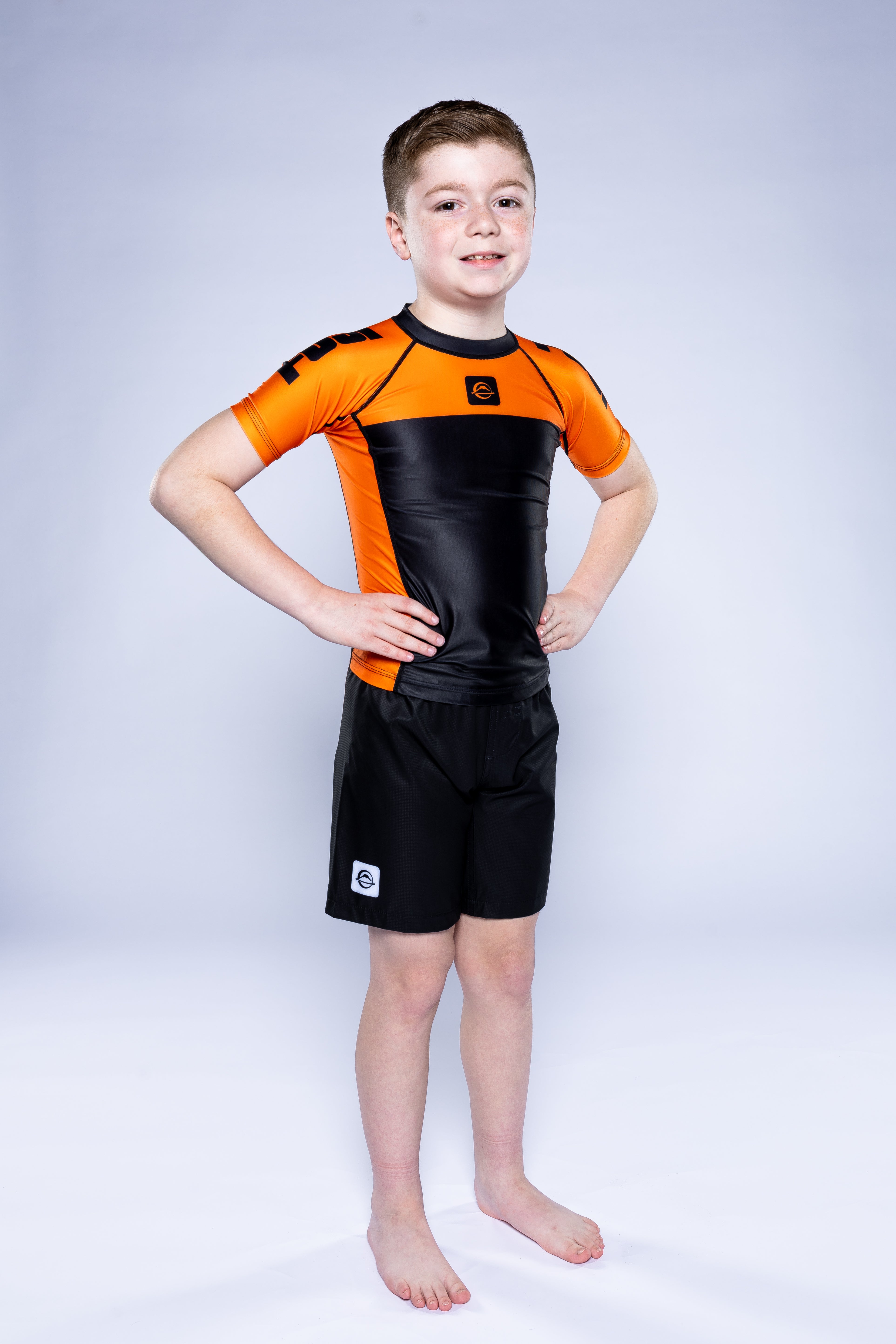 A young boy stands barefoot with hands on hips, smiling at the camera in a Kids Competition Ranked Rashguard Orange and black shorts against a plain light background.
