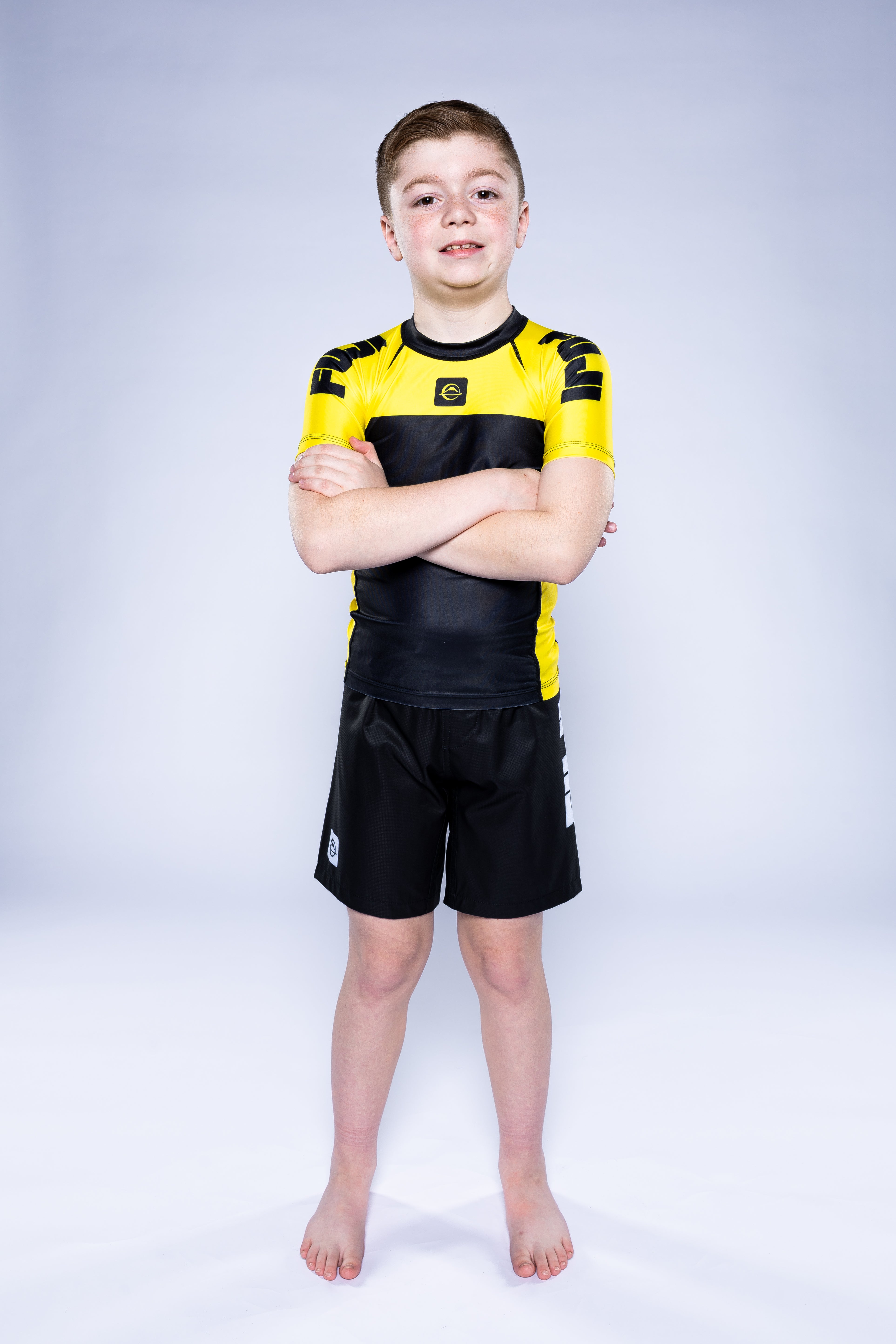 A young boy with short hair stands barefoot on a white background, smiling with his arms crossed. He is wearing the Kids Competition Ranked Rashguard Yellow and matching shorts.