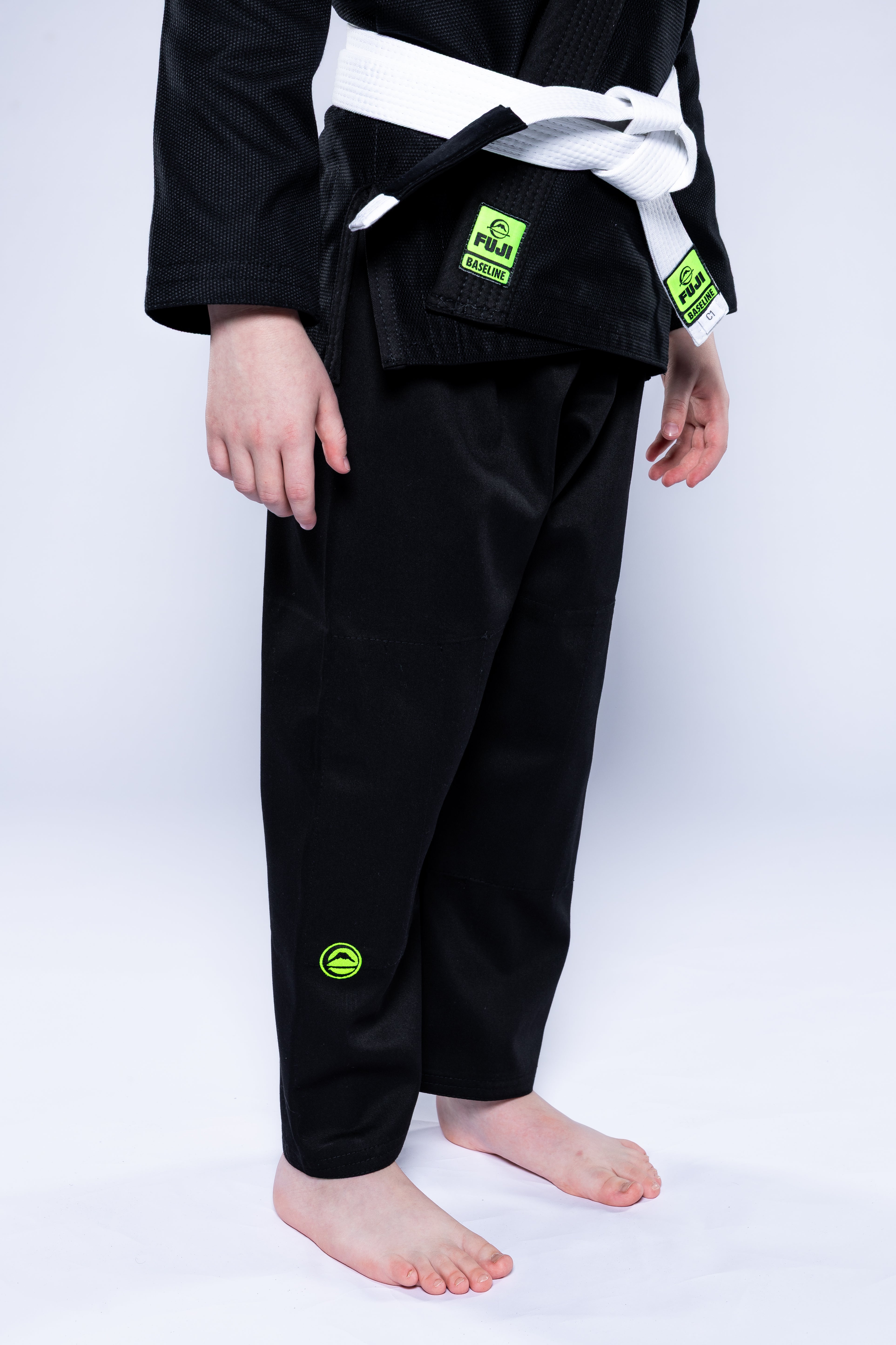 A child, barefoot, wears the Kids Baseline 2.0 Slim Fit BJJ Gi Black Green with a white belt. Only the lower half is visible against a white background, showing brand patches on the uniform.