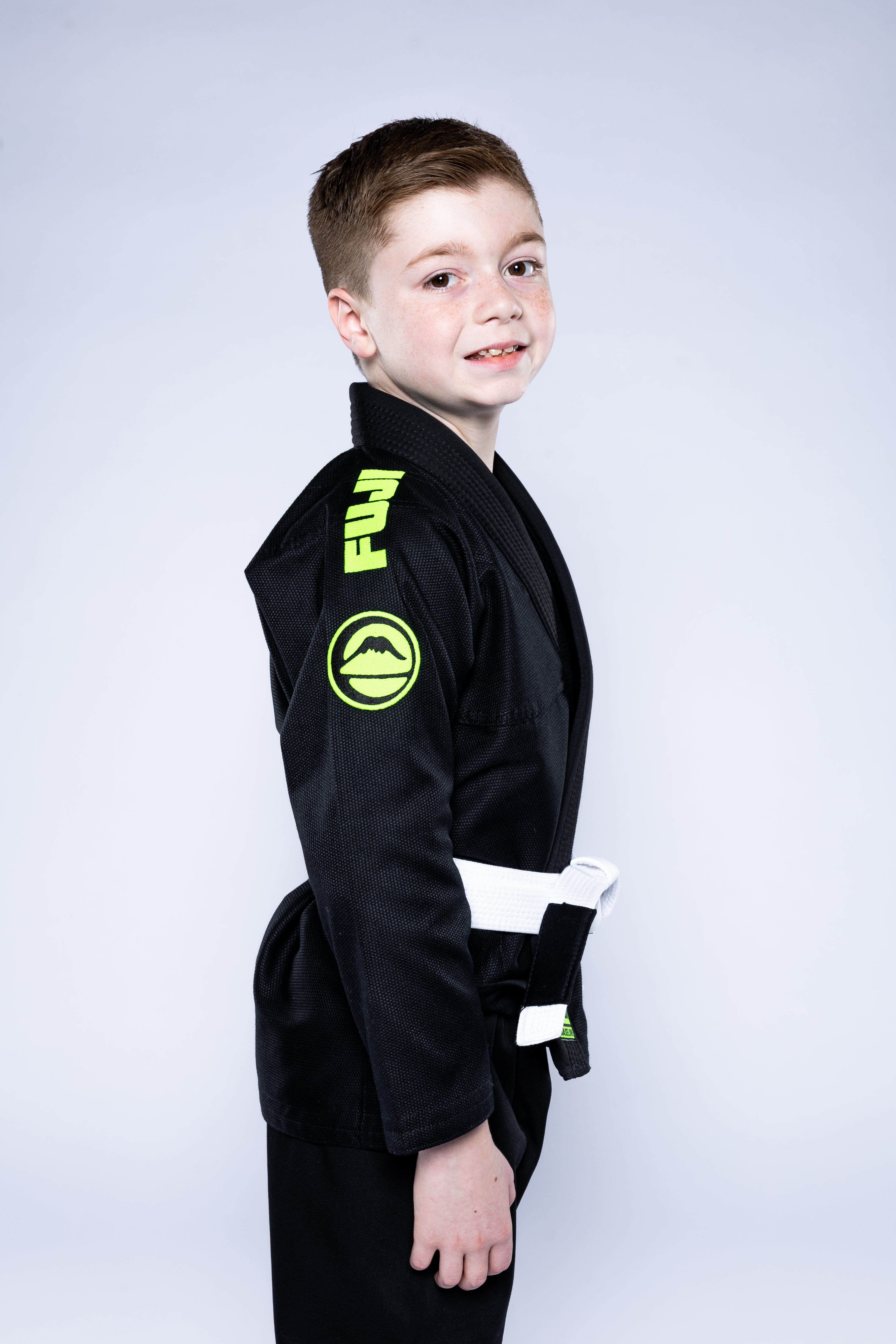 A young boy with short brown hair wears the Kids Baseline 2.0 Slim Fit BJJ Gi Black Green, styled with a white belt, standing sideways and smiling at the camera against a plain light background.
