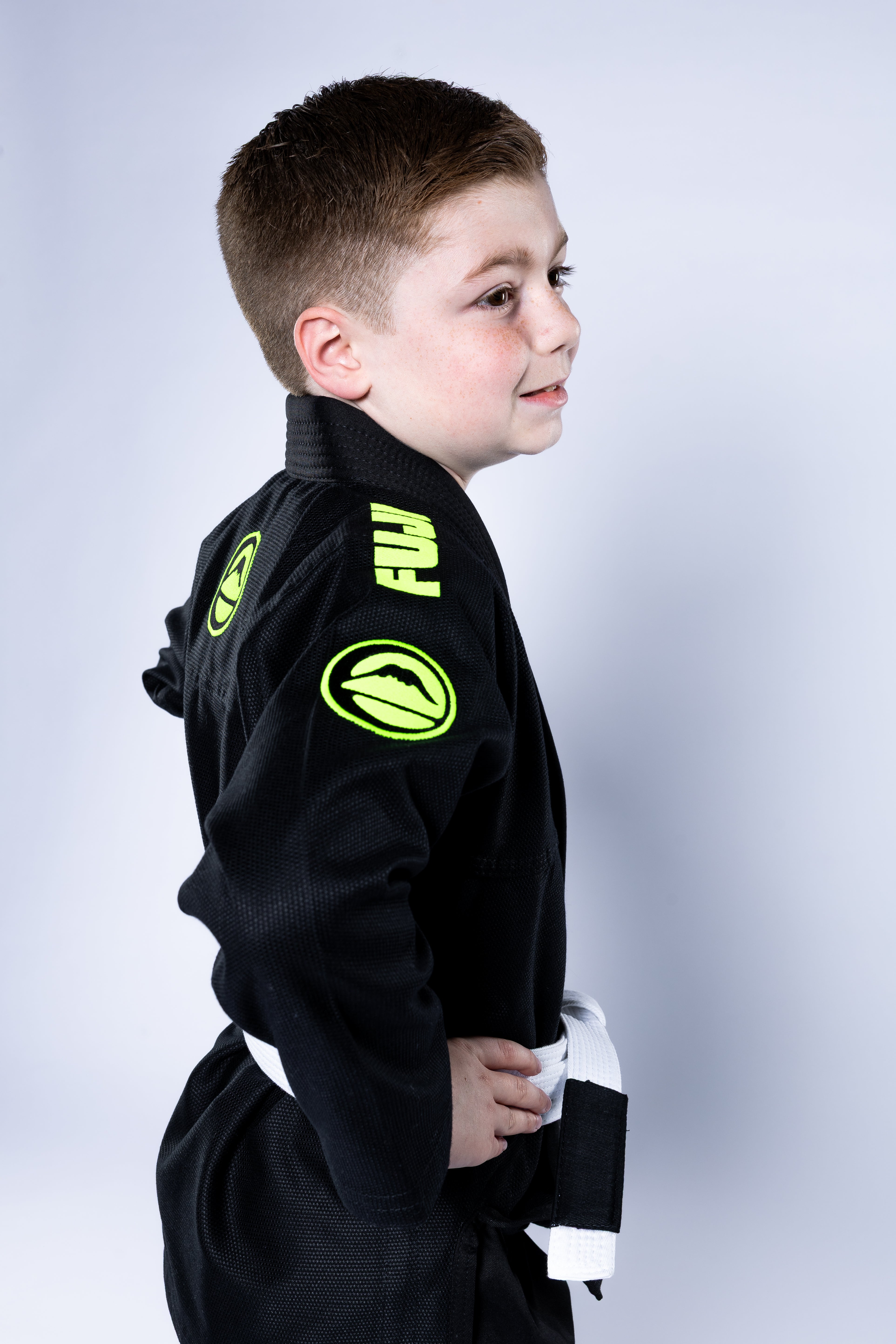 A young boy in the Kids Baseline 2.0 Slim Fit BJJ Gi Black Green with a white belt stands in profile against a plain light background, smiling with his hand on his hip.