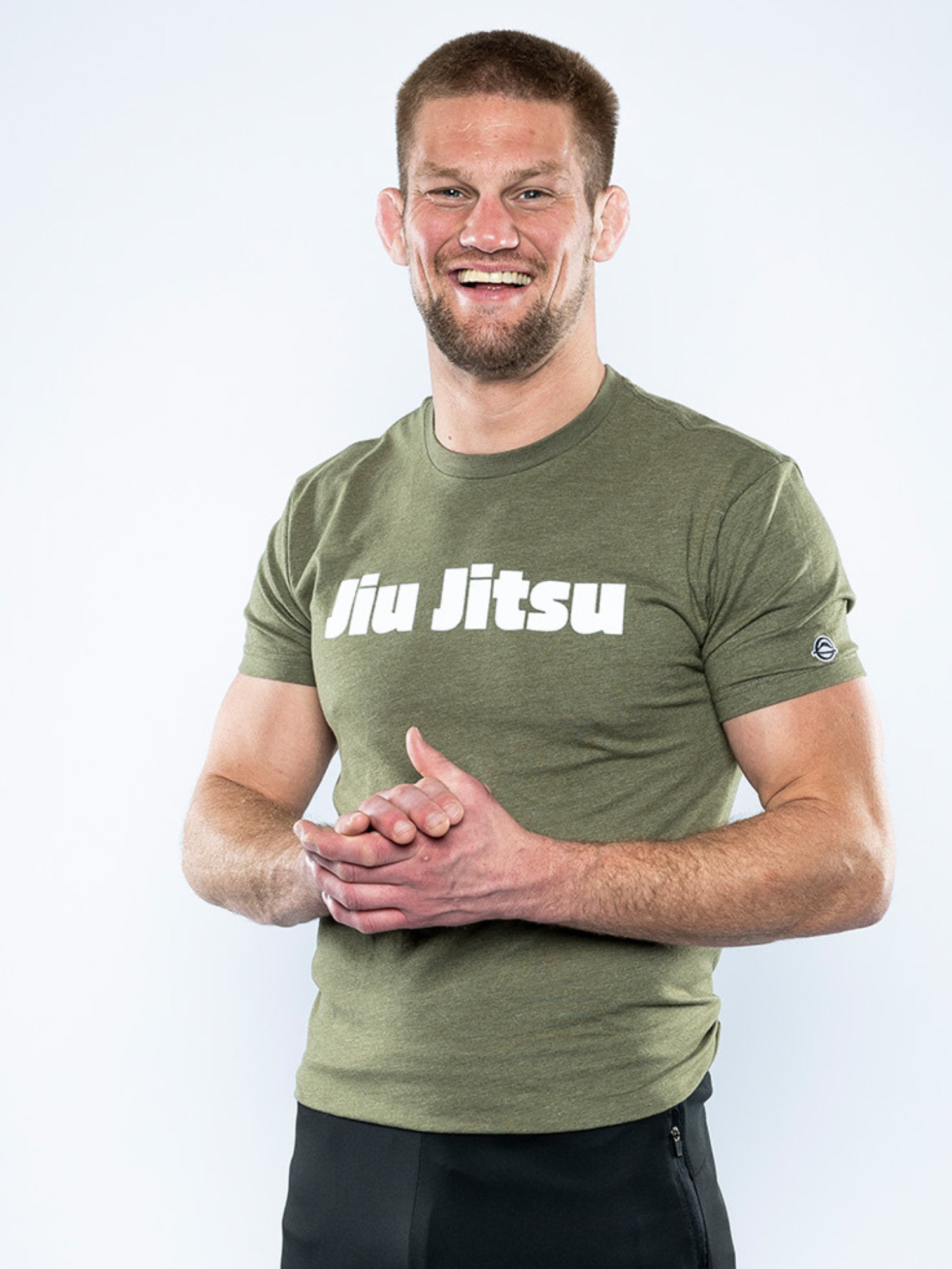 A smiling man with short hair and a beard wears the Jiu Jitsu Player T-Shirt in military green and black pants, standing against a plain white background with his hands clasped.