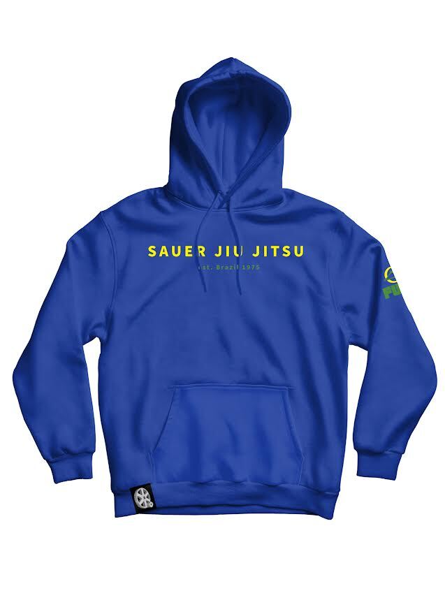 Pedro Sauer Jiu Jitsu Brazil 1975 Hoodie Blue features yellow “SAUER JIU JITSU Est. Brazil 1975” text on the front, a green logo on the left sleeve, and a black-and-white logo tag at the bottom left of the kangaroo pocket.