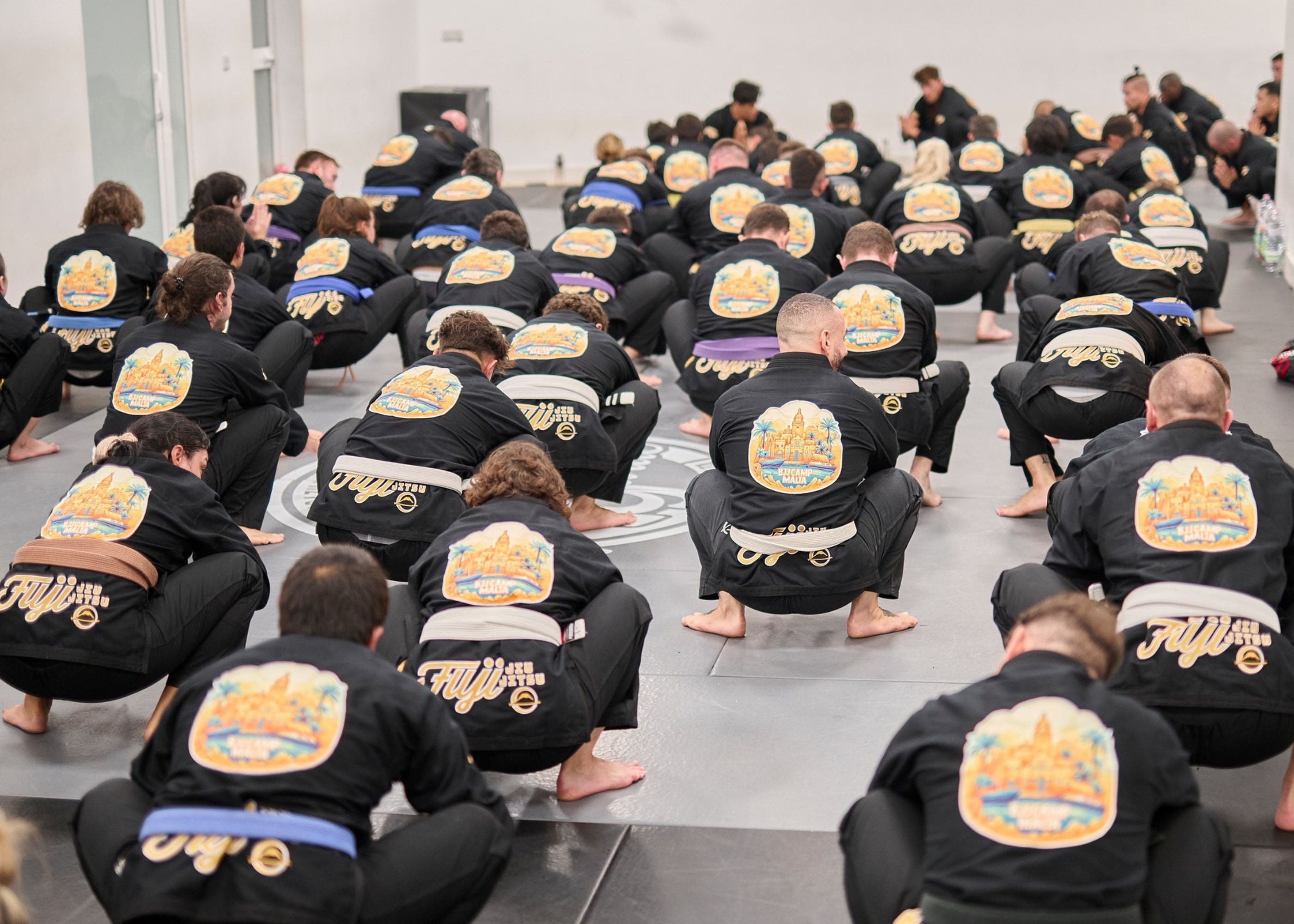 A large group of people in black martial arts uniforms with colorful patches sit in rows on a gym mat, facing an instructor at the front of a bright, spacious room.