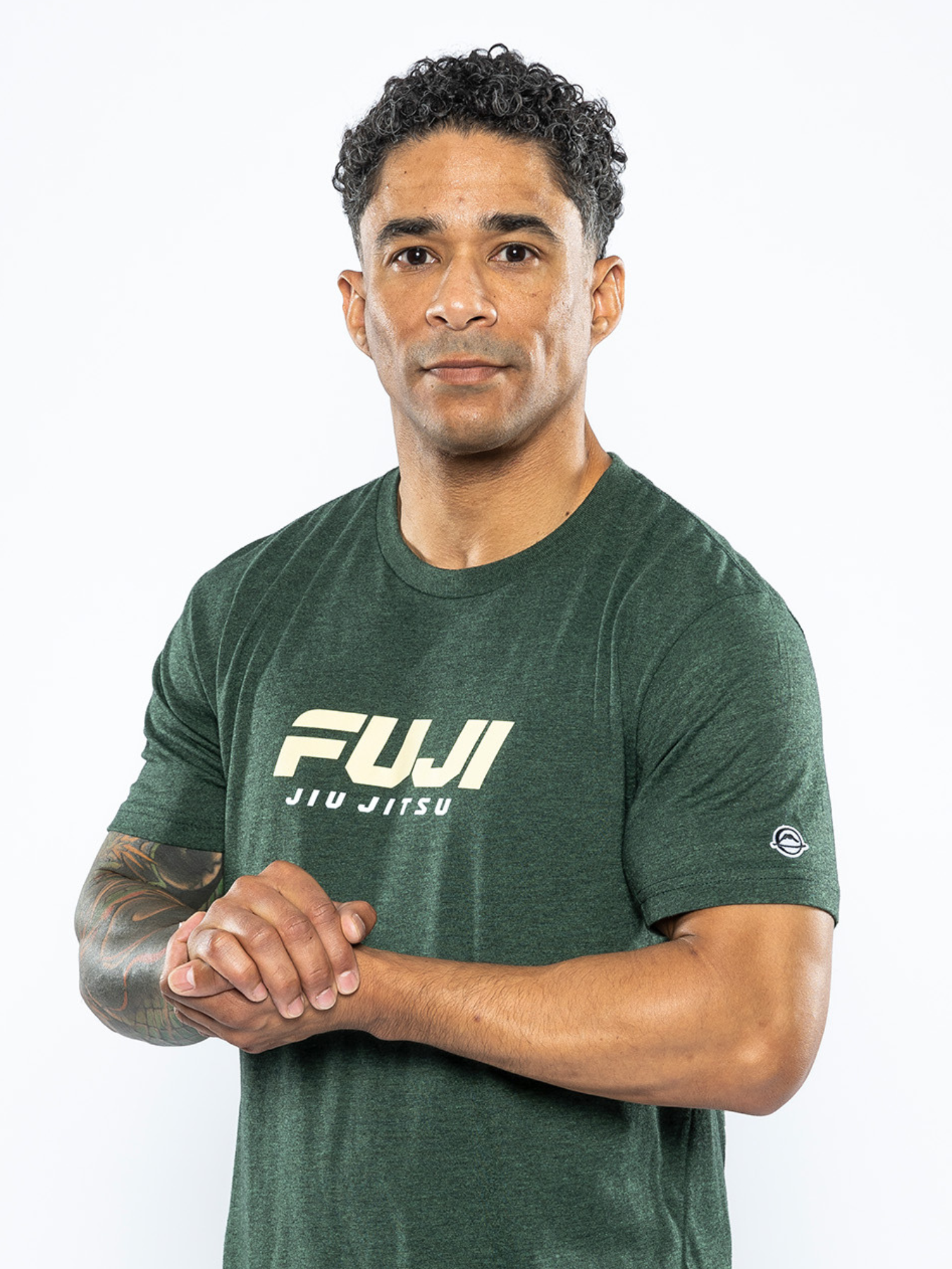A man with short curly hair stands against a white background, wearing the Apex T-Shirt in Forest Green. He has a tattoo on his left forearm and holds his hands together before him, looking at the camera.