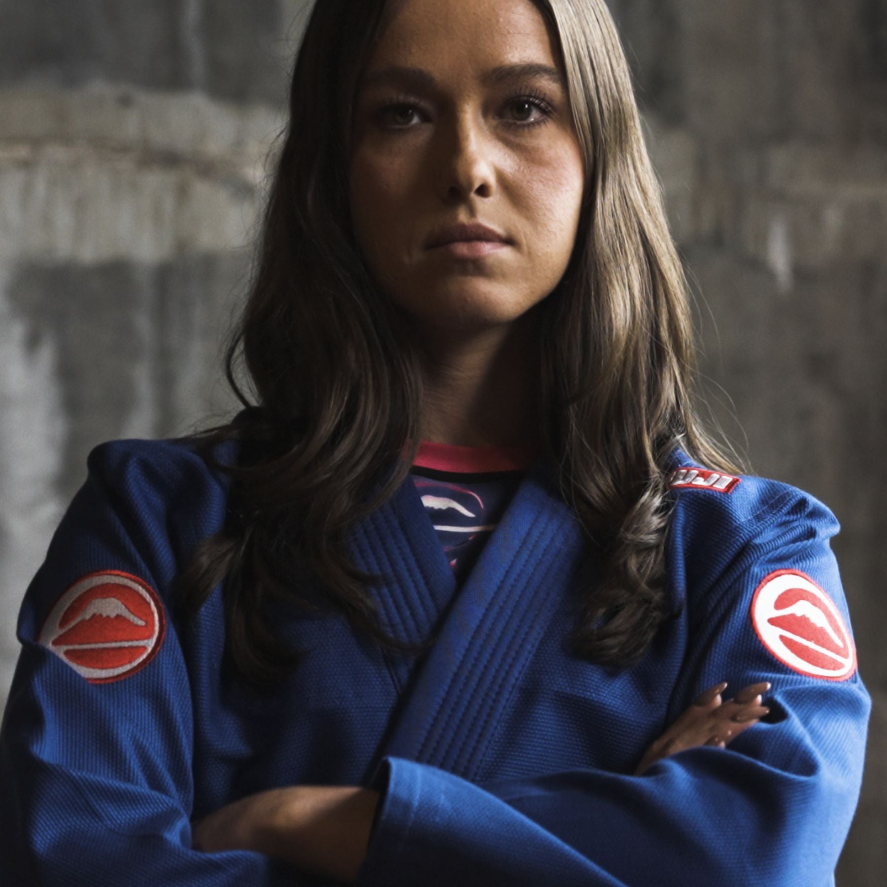 A woman with long brown hair wears a blue martial arts gi with red patches on the sleeves, standing with her arms crossed and looking confidently at the camera against a dark, textured background.
