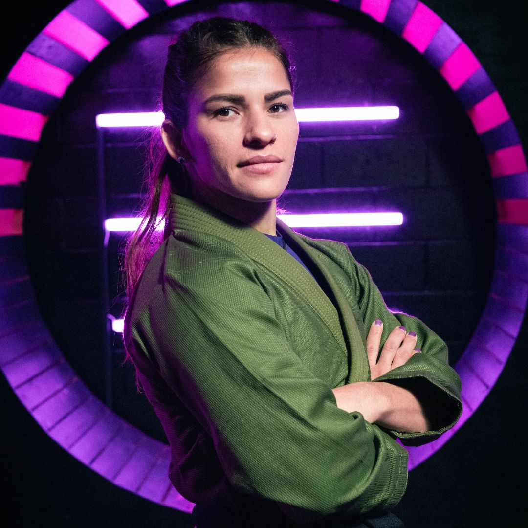 A woman in a green martial arts gi stands confidently with arms crossed. She is in front of a circular backdrop illuminated with purple and pink neon lights.