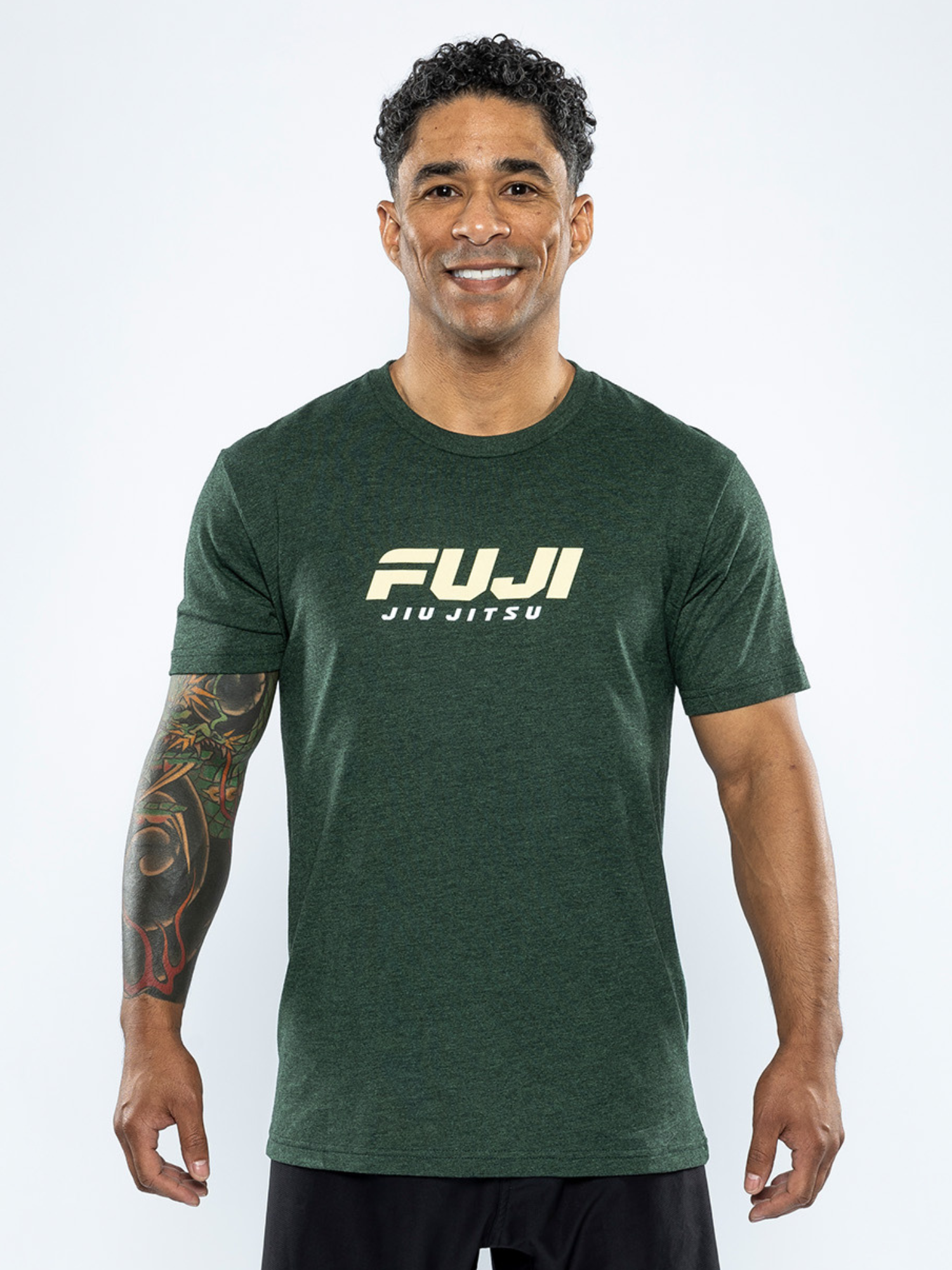 A man with curly hair and a tattoo sleeve on his right arm smiles against a plain light background, wearing the Apex T-Shirt in Forest Green.