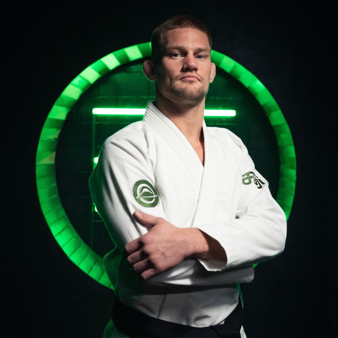 A man in a white martial arts gi poses confidently with arms crossed, standing in front of a glowing green circular light on a dark background.
