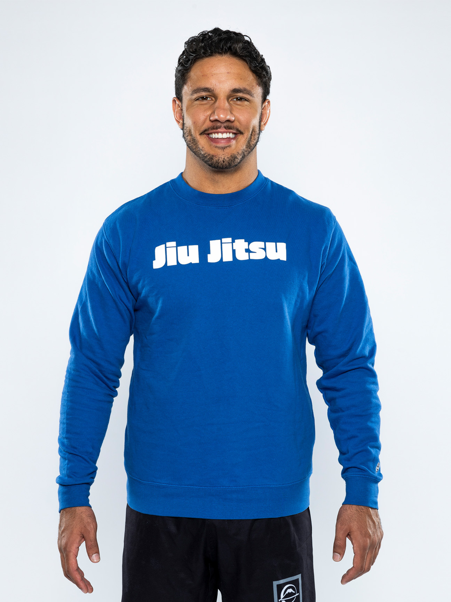 A smiling man with short curly hair wears the Jiu Jitsu Player Crewneck Blue with "Jiu Jitsu" in white letters and black shorts, standing against a plain white background.