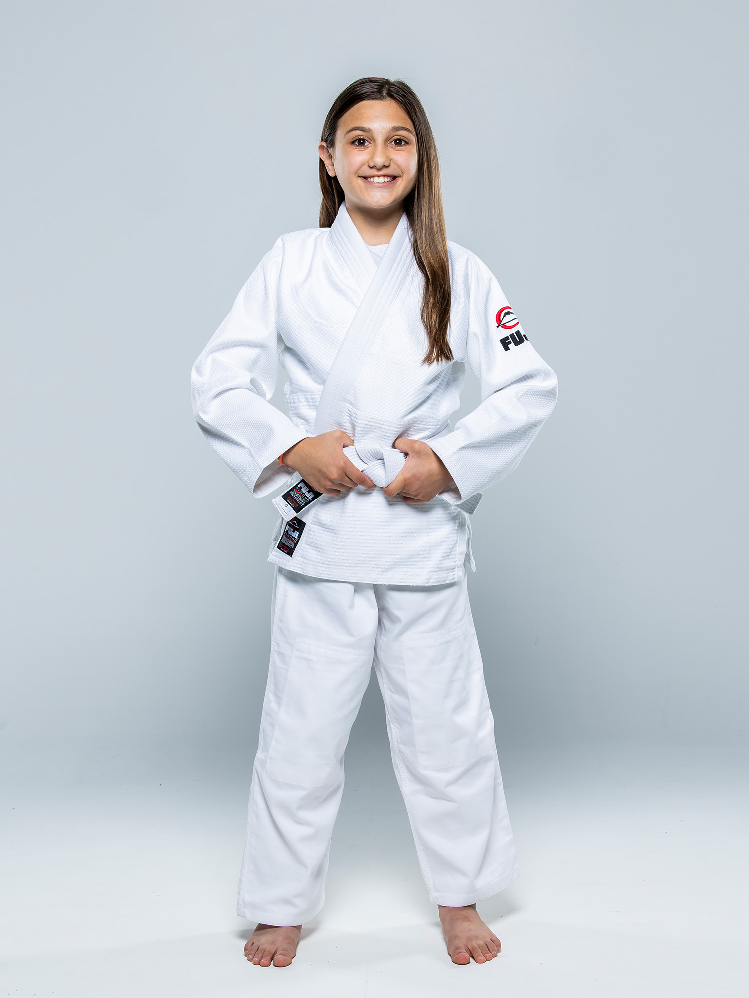 A smiling young girl stands barefoot on a plain light background, wearing the Single Weave Judo Gi White and holding the ends of her belt with both hands.