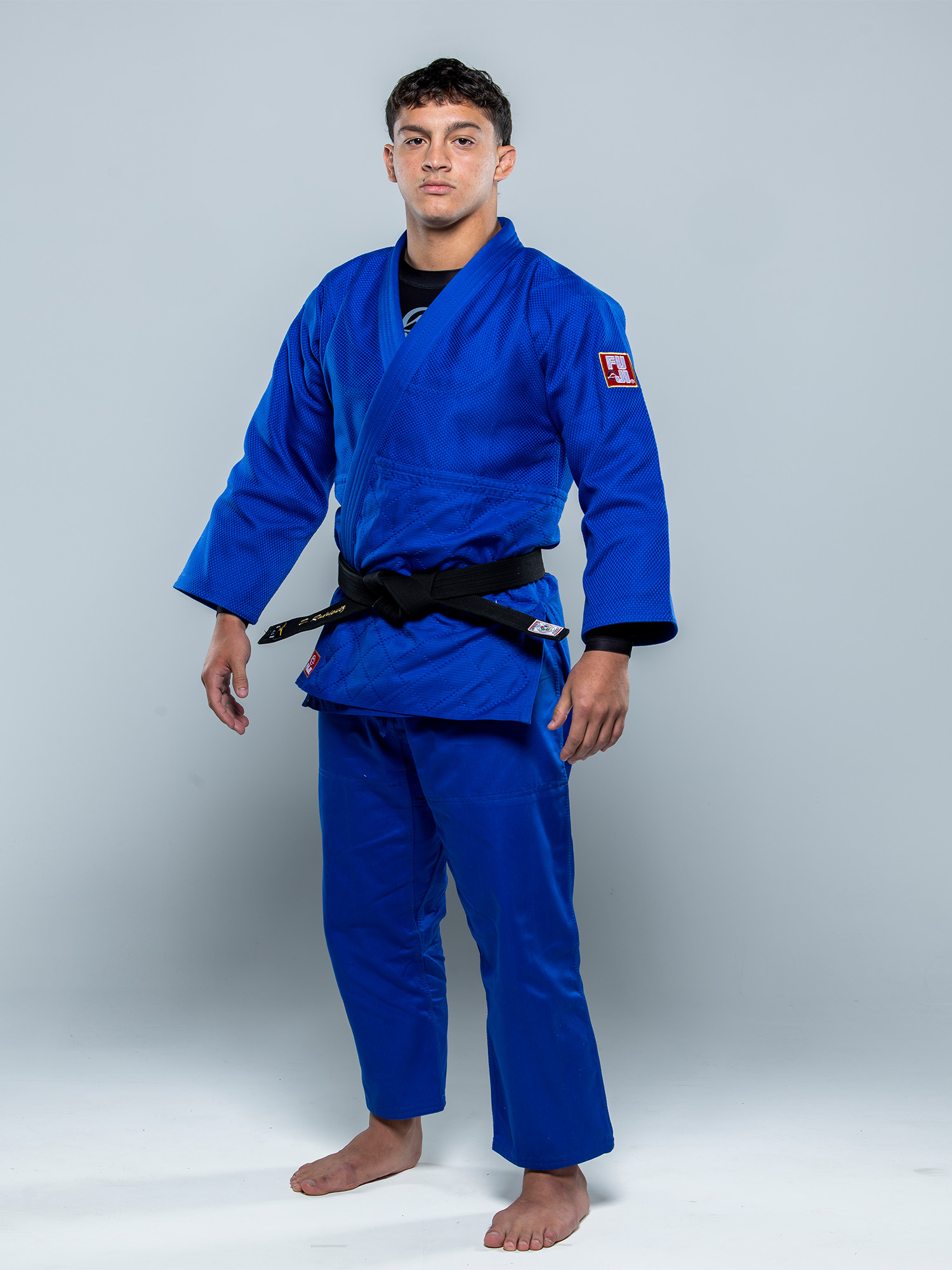 A young man stands barefoot, facing the camera against a plain light gray background, wearing the Euro Competition Judo Gi Blue with a black belt.