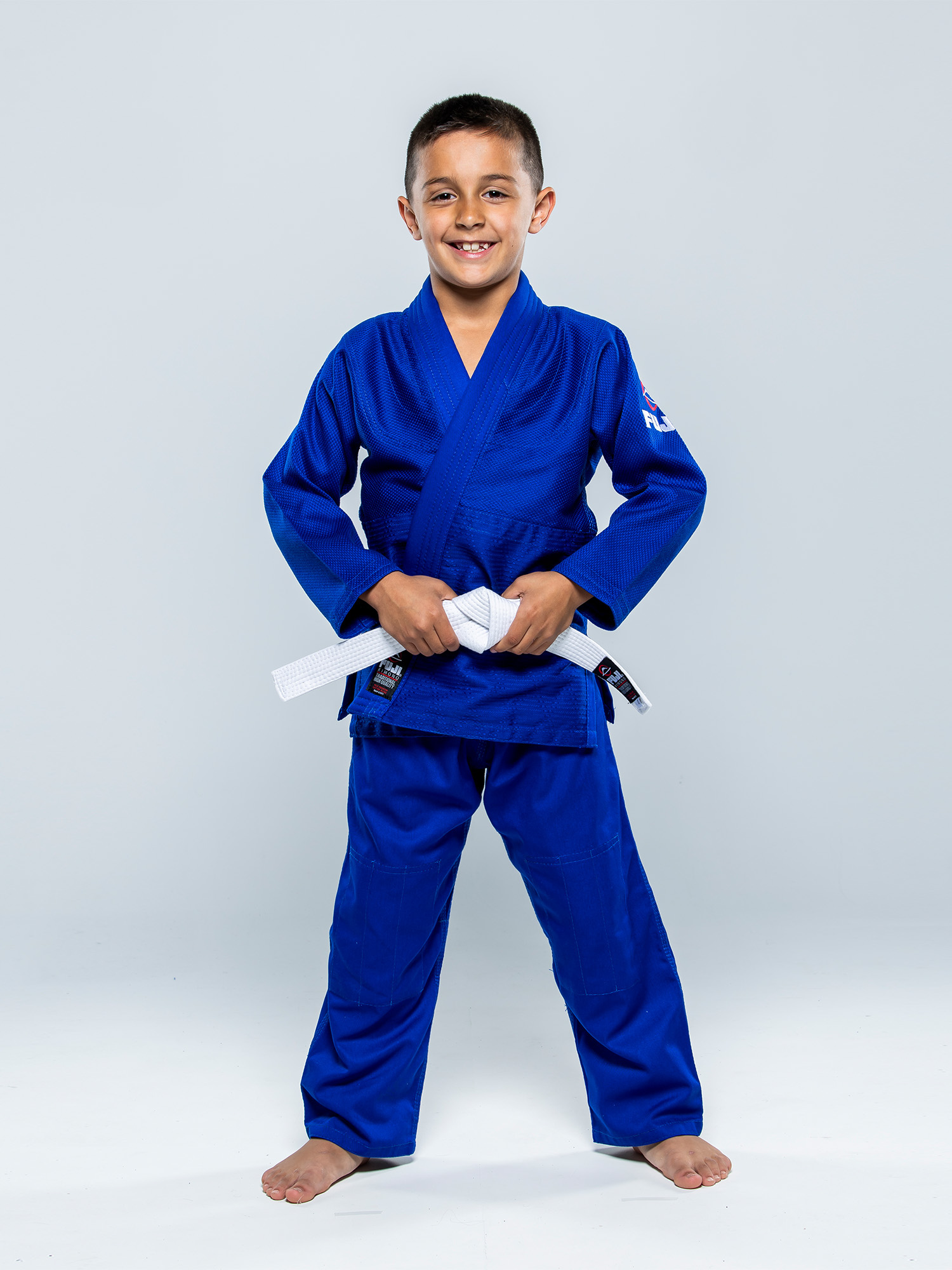 A young boy smiles barefoot on a white background, wearing a Single Weave Judo Gi Blue and holding the white belt tied around his waist.