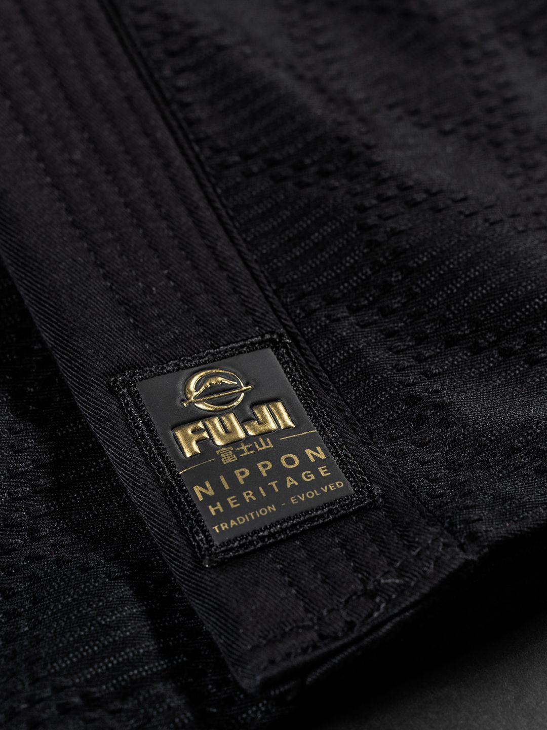 Close-up of the Womens Heritage Keisho Lightweight BJJ Gi Black, showing textured black fabric with a stitched black and gold label that reads FUJI Nippon Heritage Tradition Evolved.