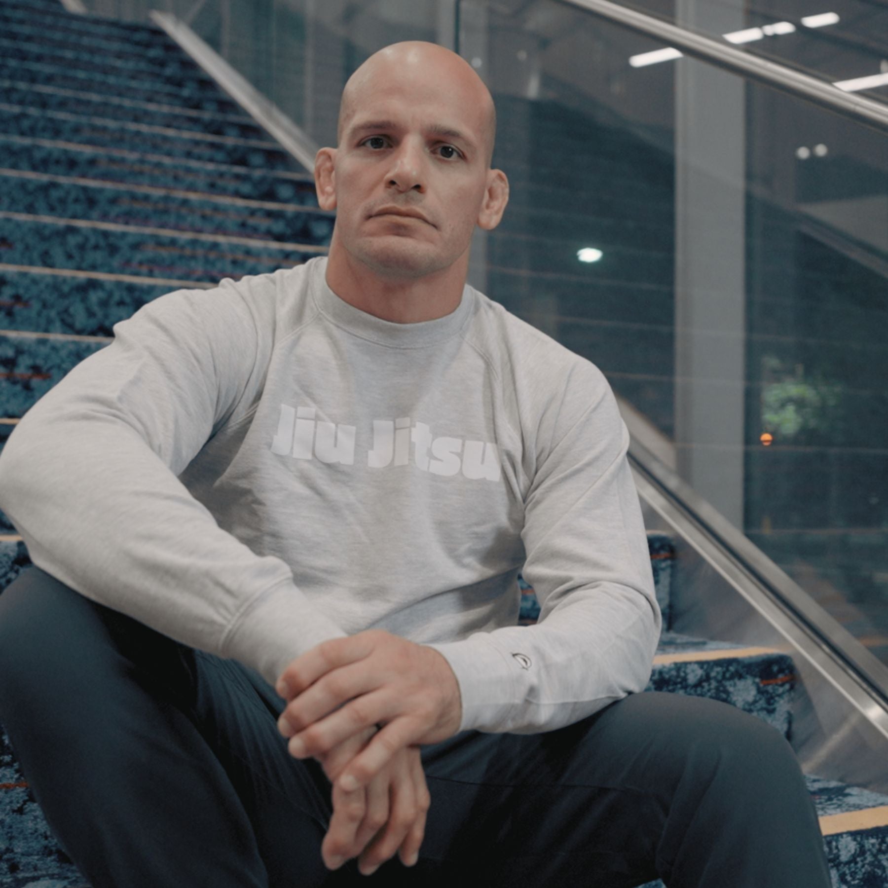 A bald man wearing a light gray Jiu Jitsu sweatshirt sits on patterned stairs indoors, looking directly at the camera with a serious expression. Glass panels and stainless steel railings are visible in the background.