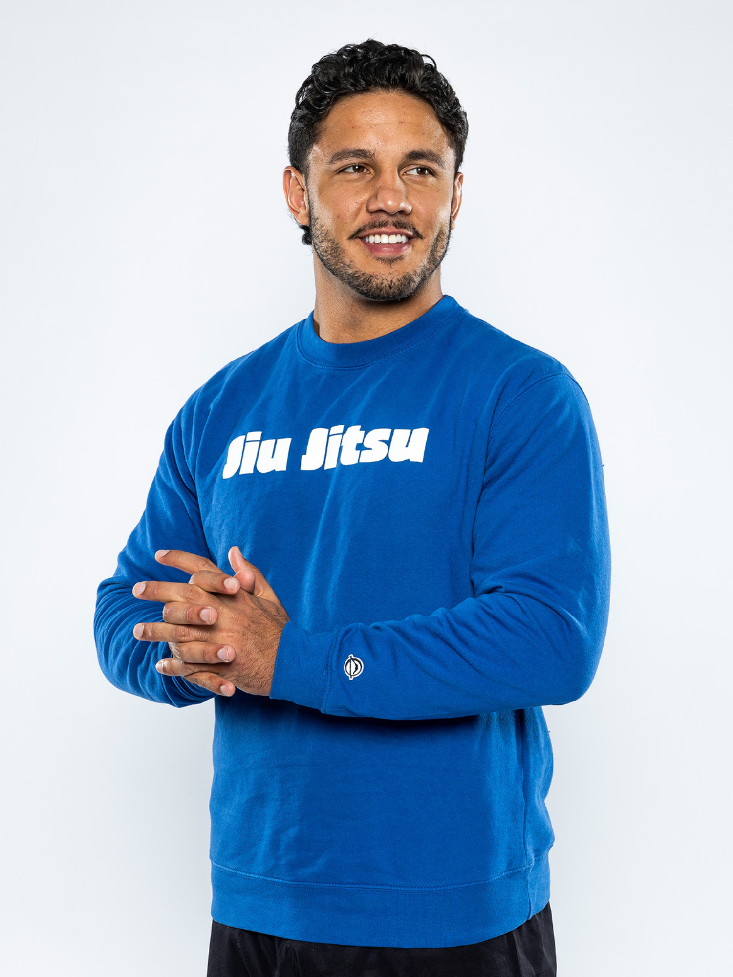 A man with short curly hair and a trimmed beard smiles while wearing the Jiu Jitsu Player Crewneck Blue, featuring "Jiu Jitsu" in white lettering on the chest, against a plain white background.