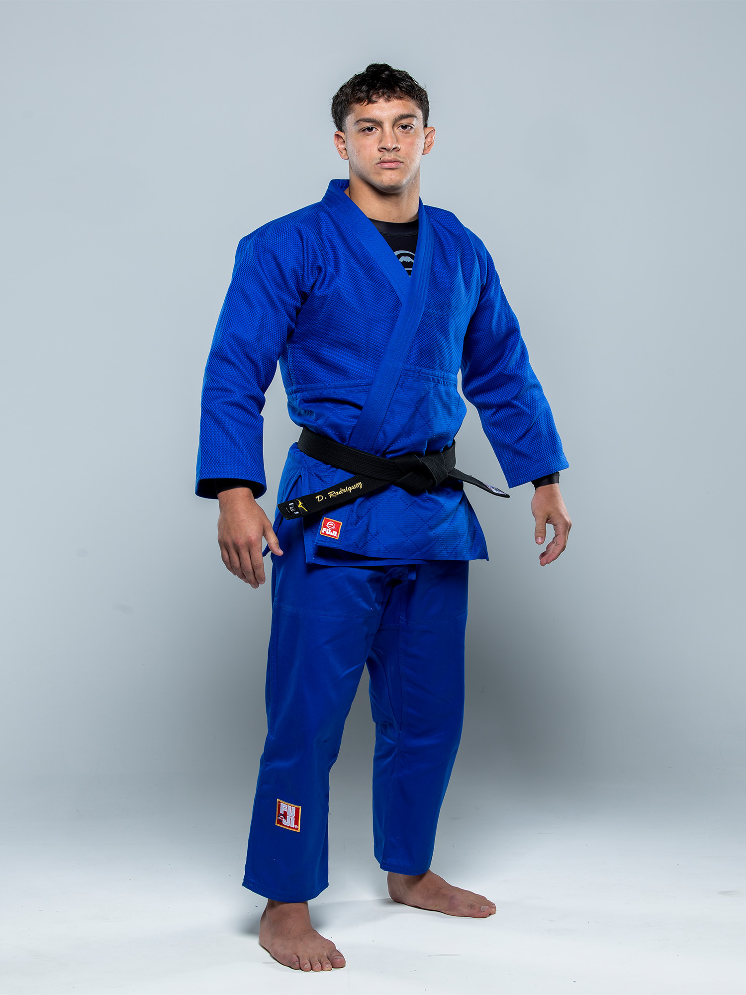 A man stands barefoot on a plain background, facing forward and wearing the Euro Competition Judo Gi Blue with a black belt. His arms are relaxed at his sides, and he has a serious expression.