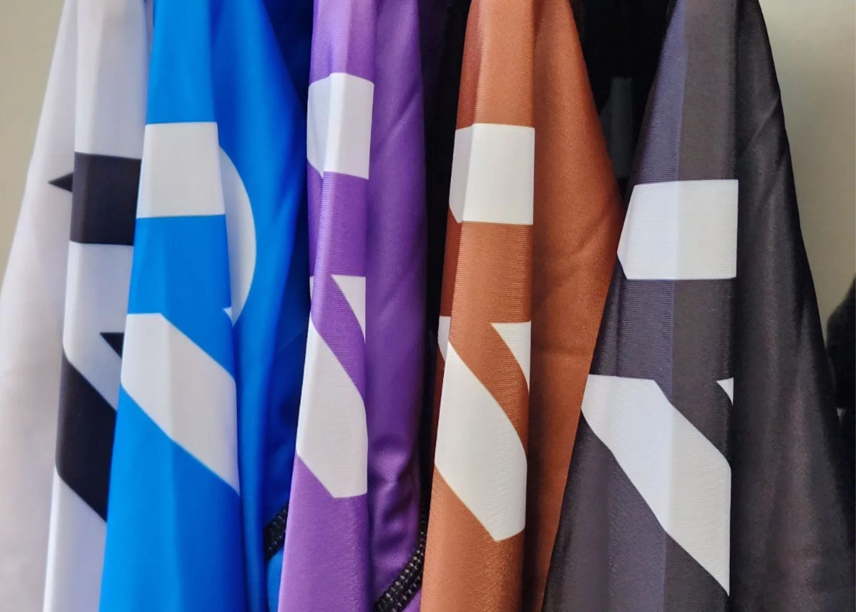 Five capes in white, blue, purple, brown, and black hang side by side. Each cape has a bold, white geometric design. The colors are vibrant and the fabric has a smooth, shiny texture.