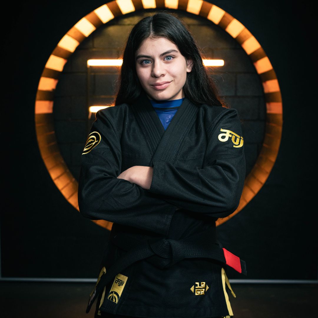 A woman with long dark hair, wearing a black martial arts gi with yellow details, stands confidently with arms crossed in front of a circular illuminated backdrop.