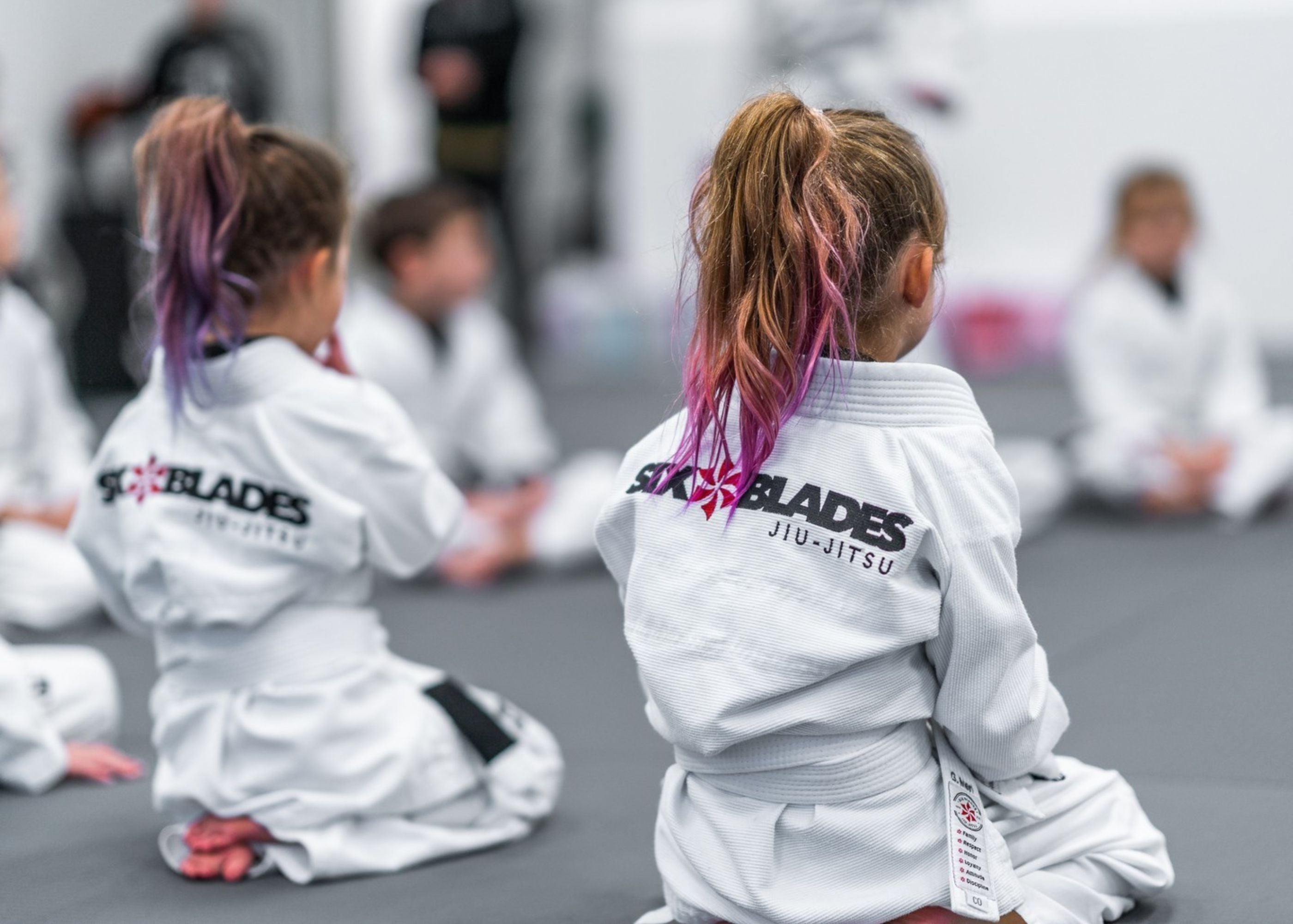 Young children wearing white jiu-jitsu uniforms with Six Blades Jiu-Jitsu on the back sit cross-legged on a mat, facing forward. Their hair is tied up, some with pink tips. The background is blurred.